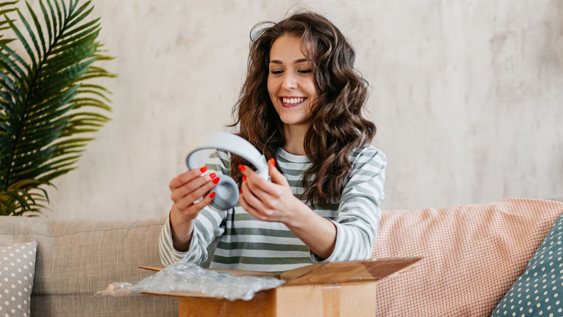 Mujer joven morena con pelo rizado abriendo una caja con auriculares en el salón de su casa