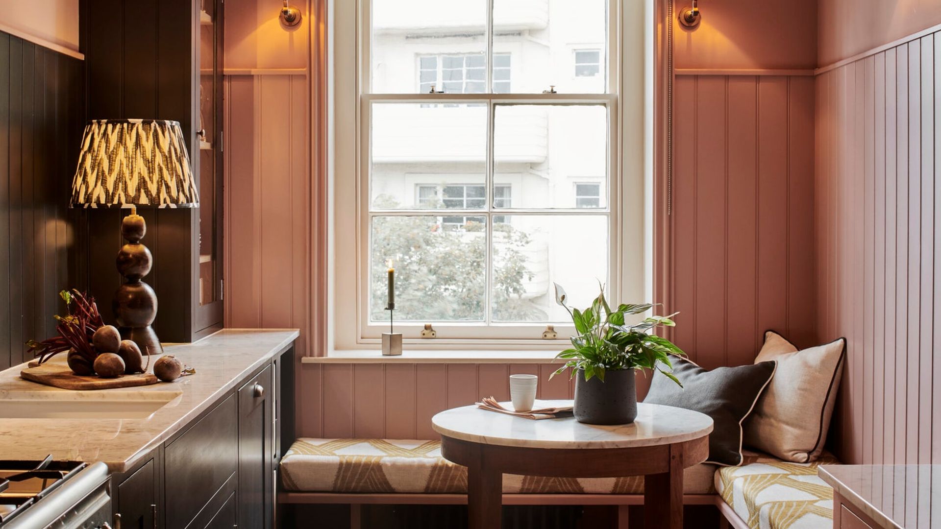 Cocina con friso de madera rosa pastel en las paredes, zona de office con banco y mesita bajo la ventana, apliques de pared, encimera blanca y muebles negros 