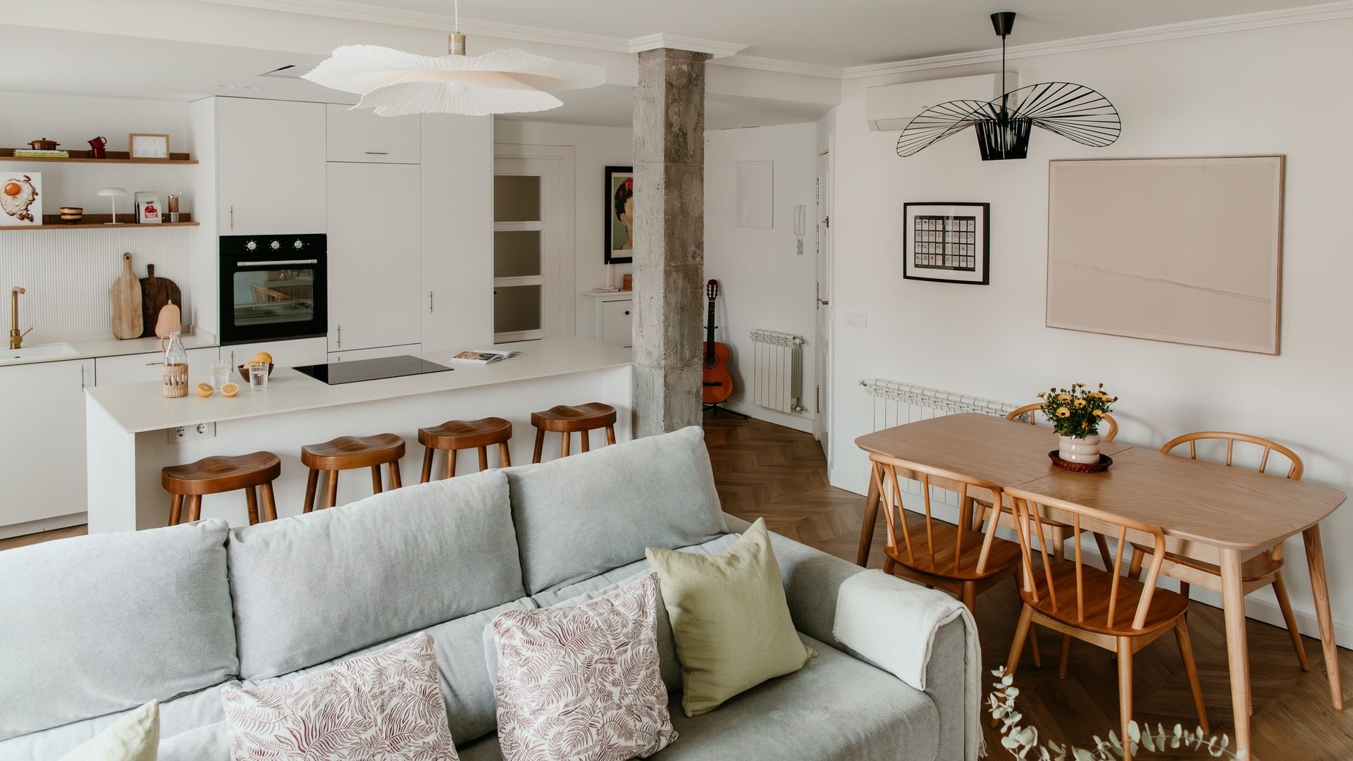 Salón comedor con cocina con isla blanca, mesa de madera, sofá gris, lámpara de techo Pamela, suelo de madera, columna cemento