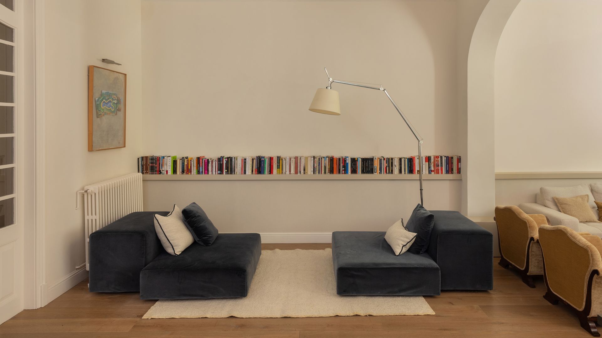 Rincón de lectura o de biblioteca con balda de pared a pared con libros, dos daybed de color azul, lámpara de pie, molduras en el techo, radiador antiguo, cuadro en la pared con aplique, alfombra