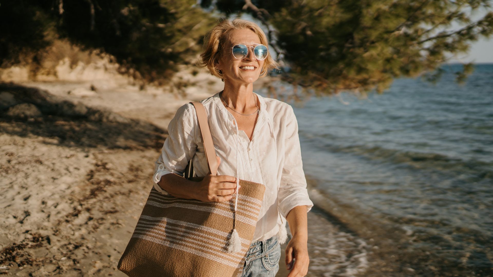 Mujer madura con pelo corto, camisa blanca y bolso de rafia en la playa