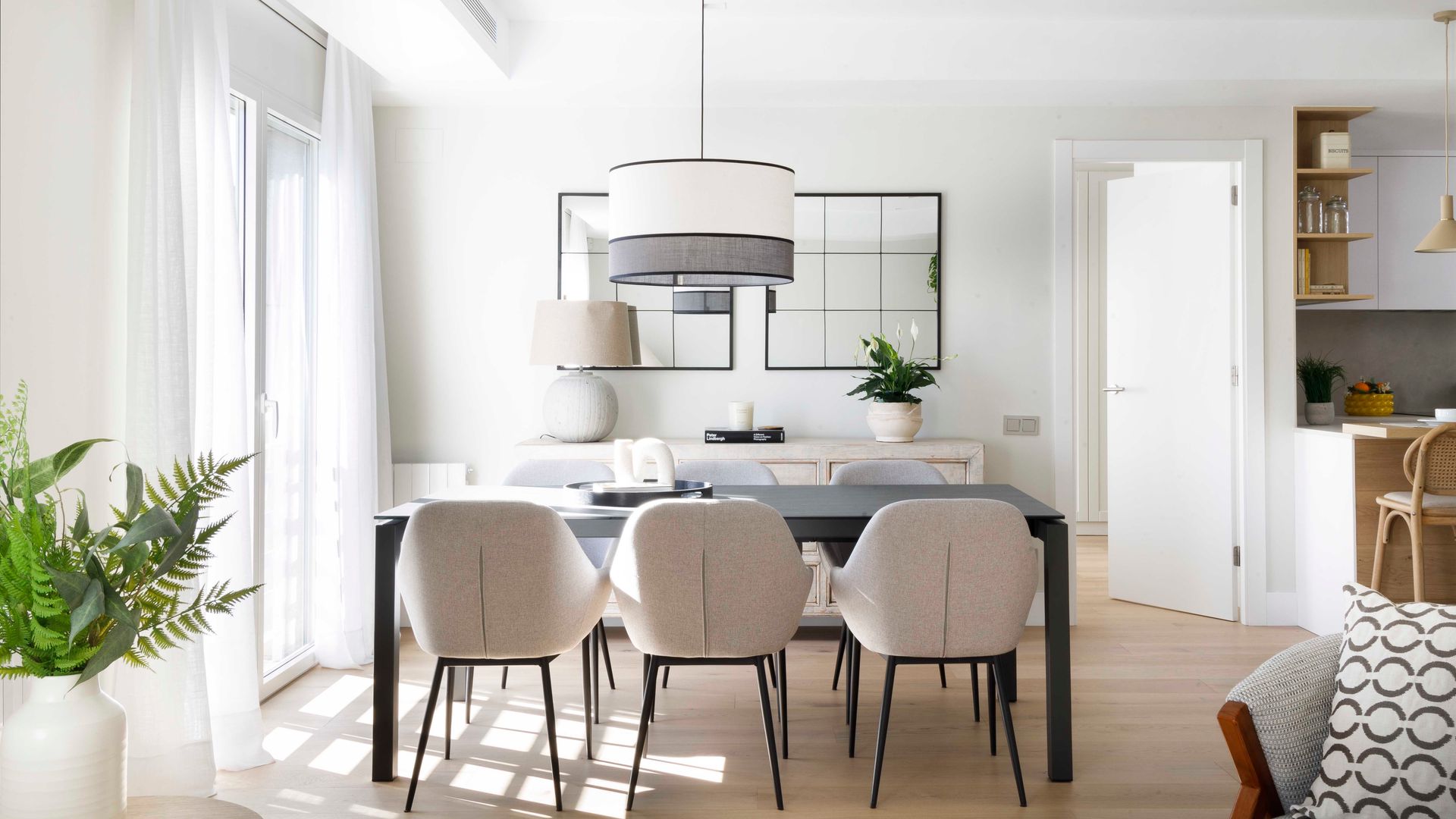 Comedor con visillos blancos, espejos, mesa de comedor rectangular negra, sillas de comedor tapizadas, suelo de madera, lámpara de techo con pantalla de tela