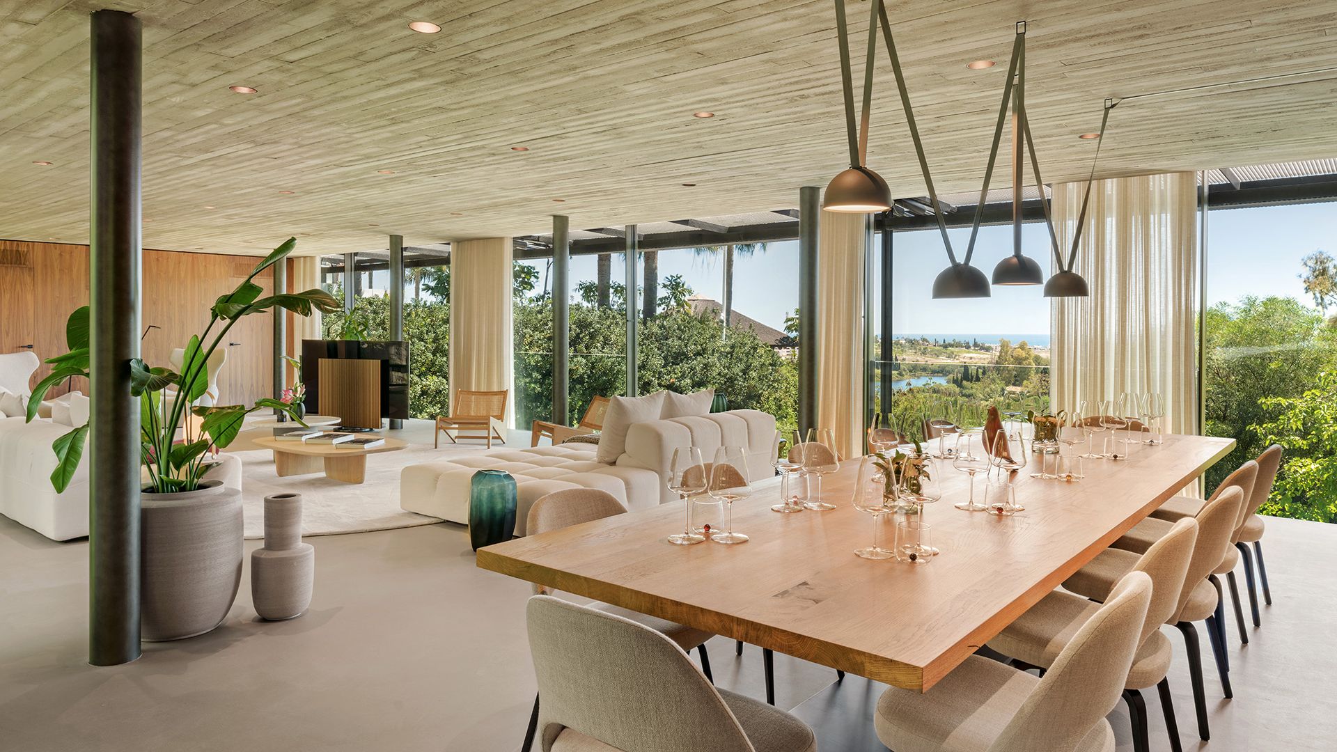 Salón comedor con mesa de madera XL, columnas, techo de hormigón, tabiques de cristal, cortinas beiges y lámparas de techo sobre la mesa