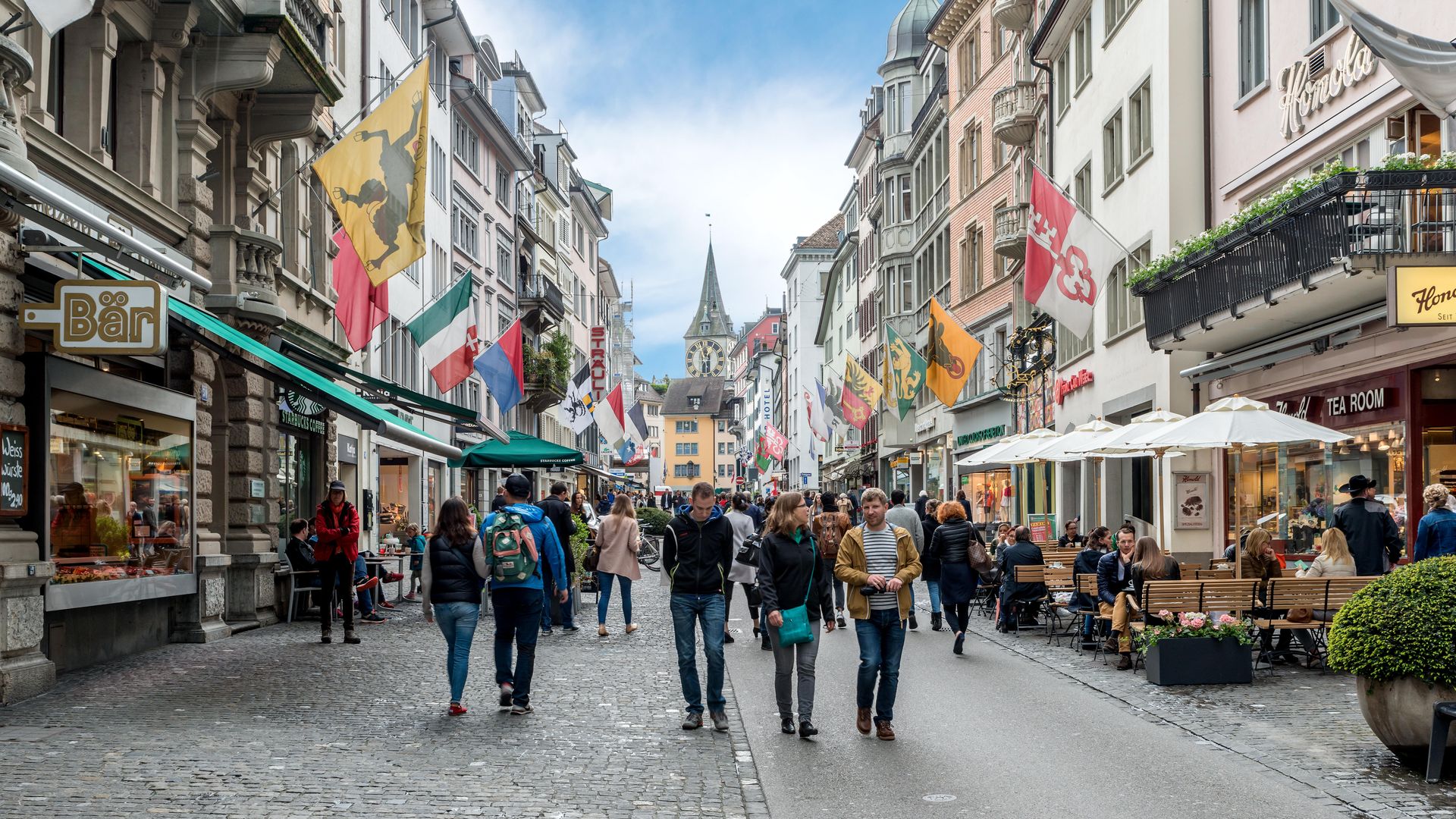 calle comercial Rennweg, con boutiques, banderas en los antiguos edificios y la torre del reloj de la iglesia de San Pedro al fondo, Zúrich