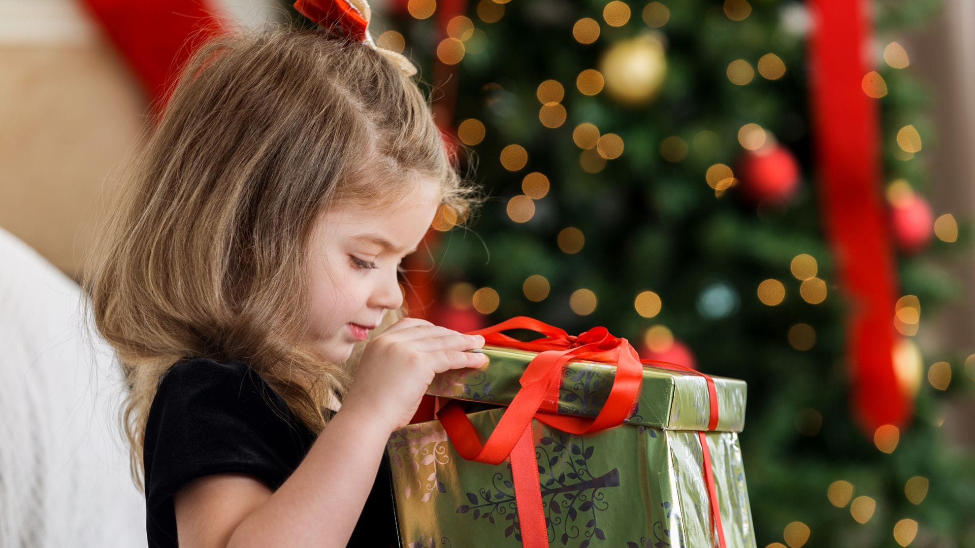 Niña abriendo una caja de regalo en rojo y verde por NAvidad, con un vestido de terciopelo negro, un árbol de Navidad detrás y mirando dentro ed la caja.