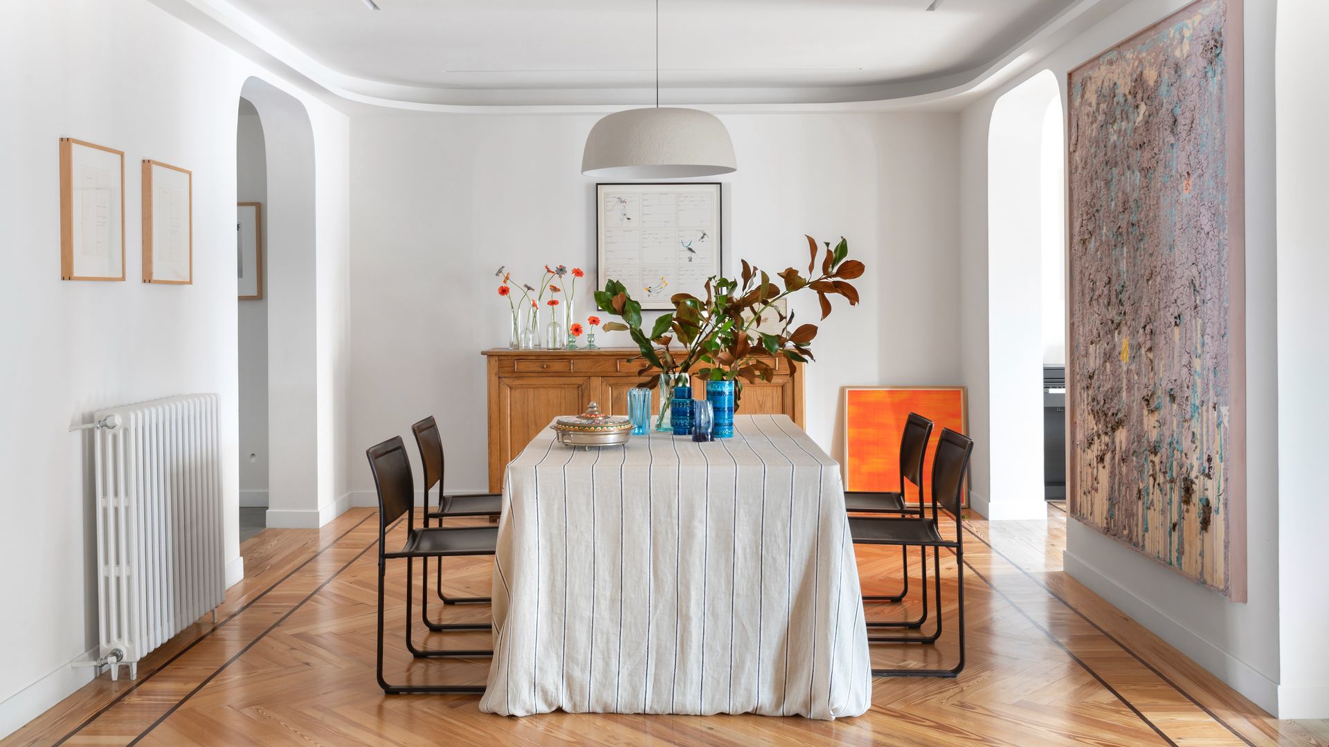 Comedor con gran mesa en el centro con mantel blanco, lámpara de techo blanca, aparador de madera, cuadro Xl en la pared, suelo de madera en espiga, radiador
