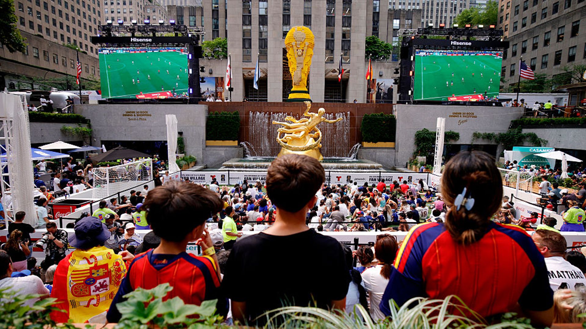 La escultura de bronce dorado de Prometeo y una réplica de 8,2 metros (27 pies) del trofeo de la Copa del Mundo, construida por el Grupo LEGO, presiden a los aficionados que siguen la semifinal del Mundial de fútbol entre Francia y España en la NYNJ World Cup 26 Fan Village, instalada en el Rockefeller Center, en Nueva York