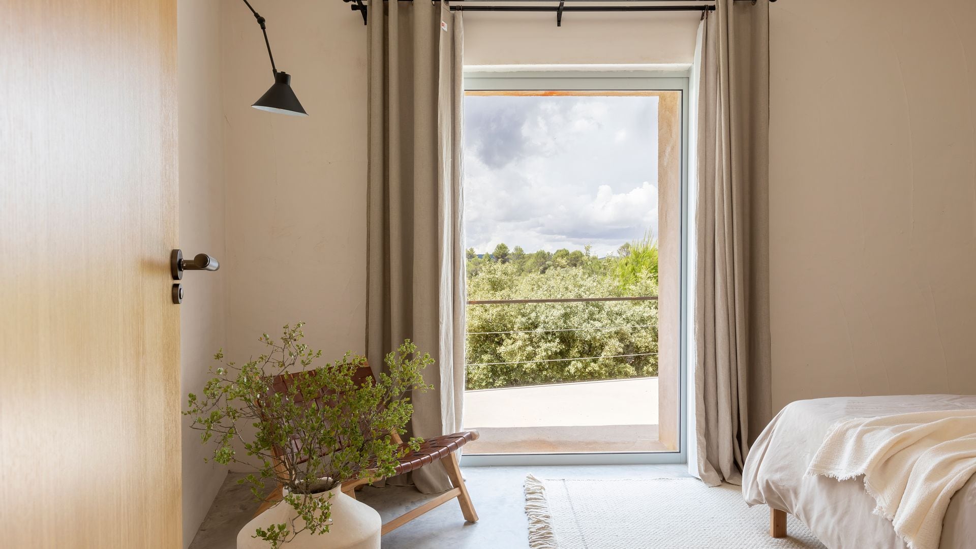 Dormitorio en tonos beiges, con grandes ventanales con cortina, alfombra, lámpara, silla de madera, jarrón