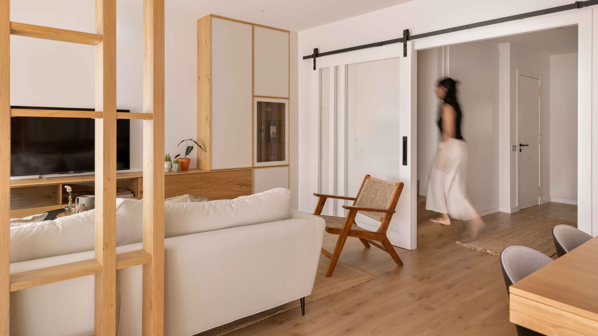 Salón de estilo japandi, con puerta corredera blanca acristalada, mujer pasando, sofá blanco, tabique de lamas de madera