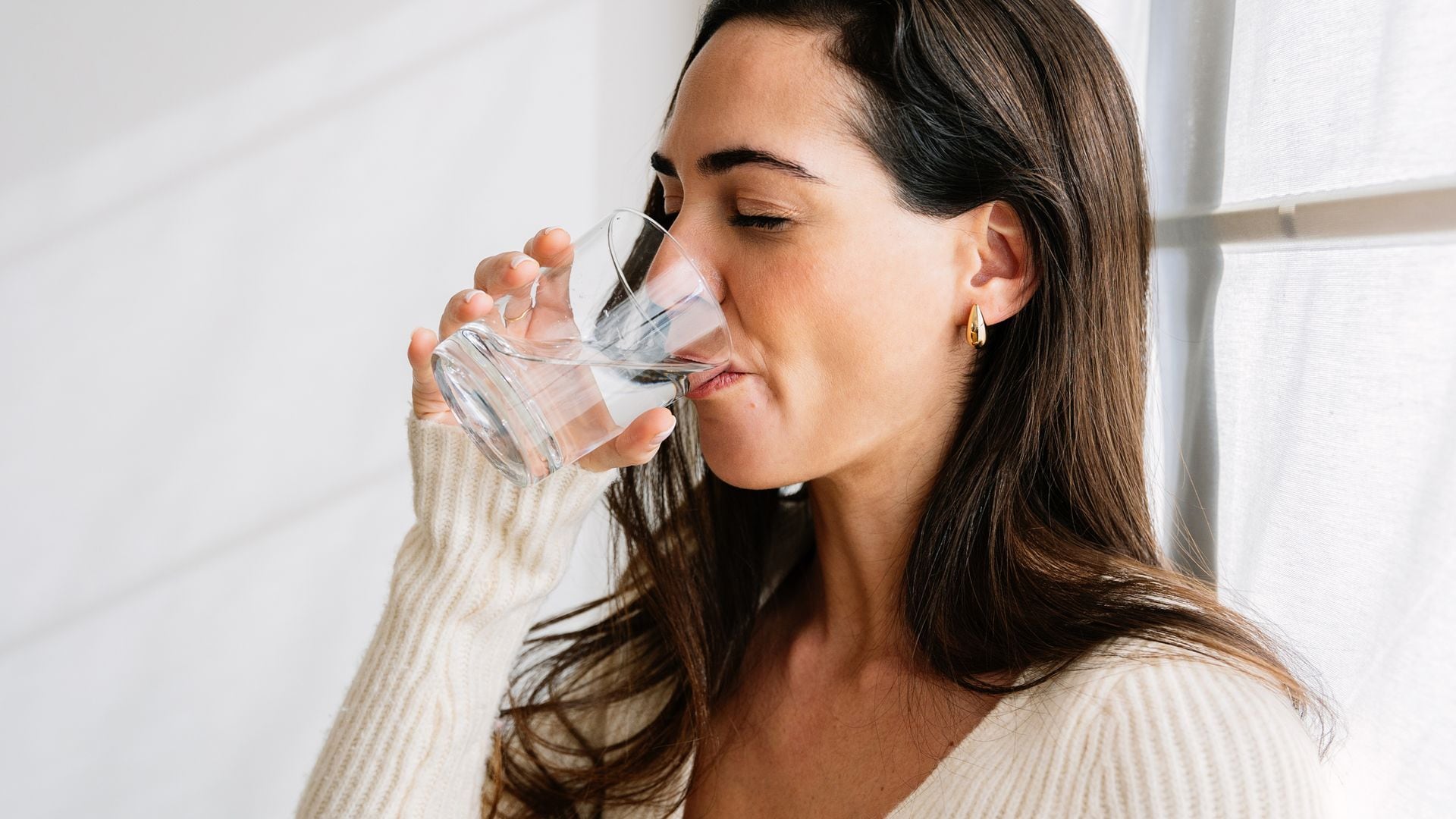 Mujer bebiendo un vaso de agua en la cocina con los ojos cerrados.