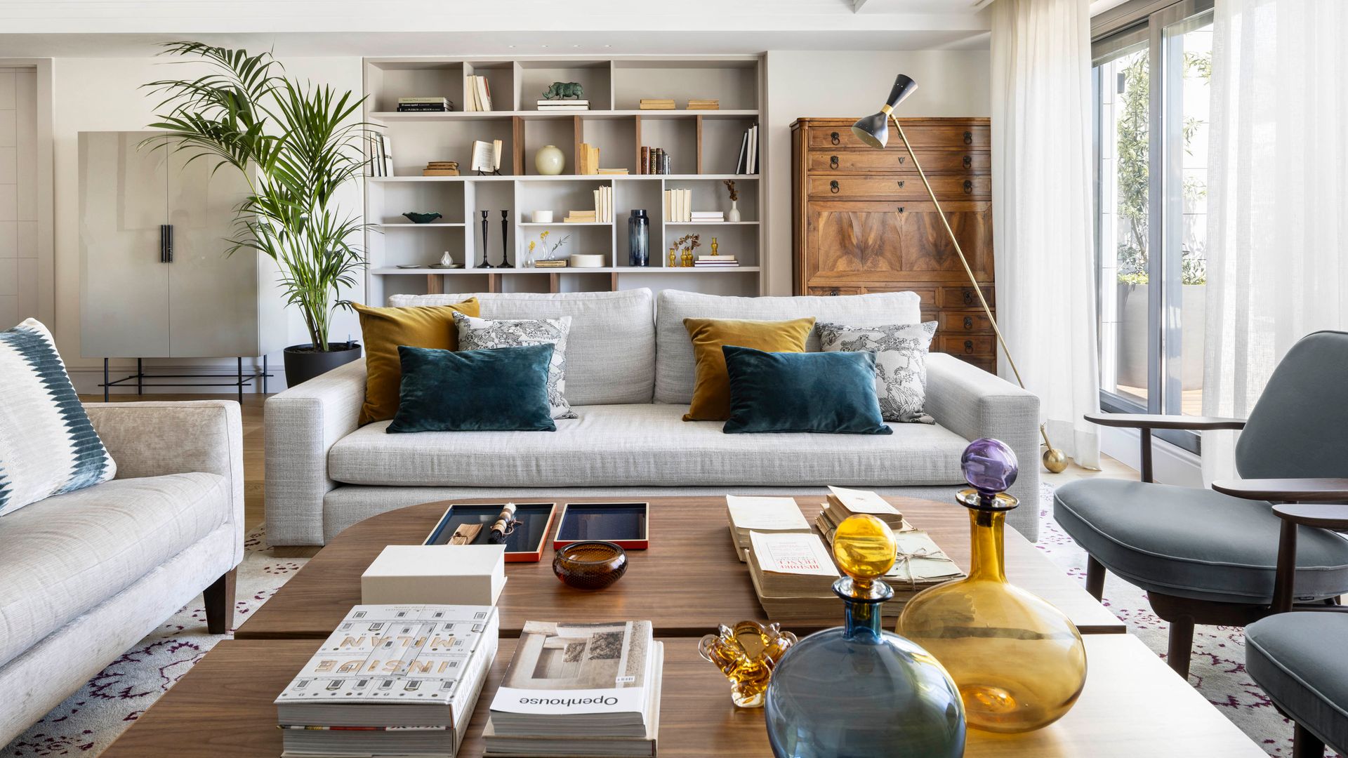 Salón con librería, escritorio antiguo de madera, sofás de color gris, mesas de centro de madera con libros, jarrones de colores y adornos, sillas grises, lámpara de pie
