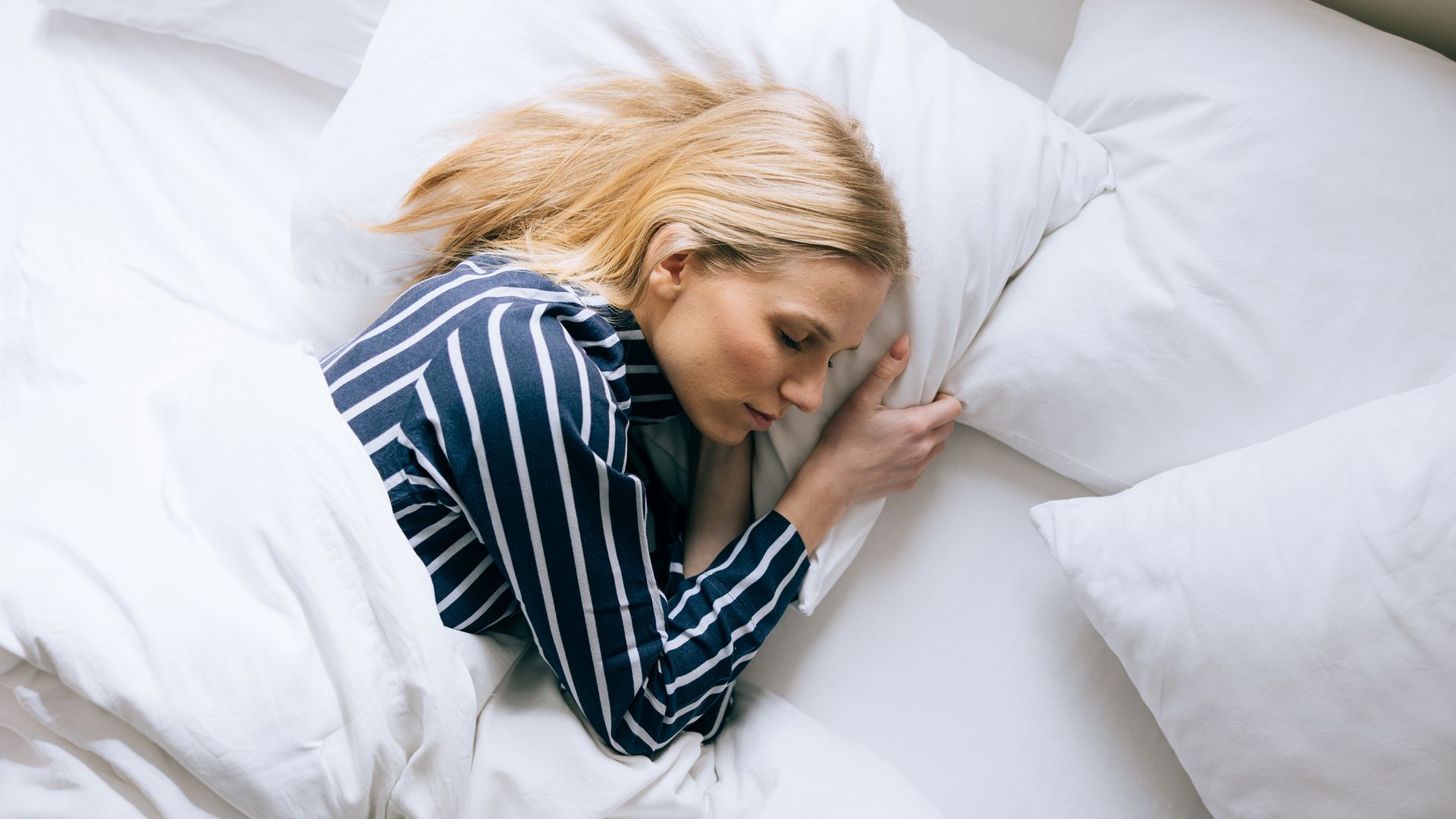 Mujer durmiendo en una cama blanca, plácidamente. Es una mujer joven rubia, con pijama en azul marino con rayas blancas.