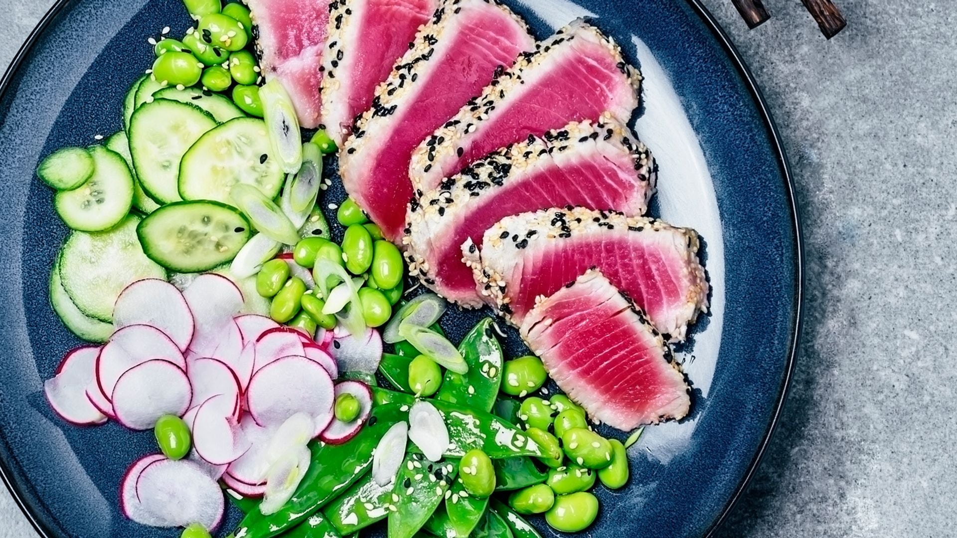 A plate of sliced tuna steak with fresh salad (radish, cucumber, edamame, and sugar snap peas) on gray background