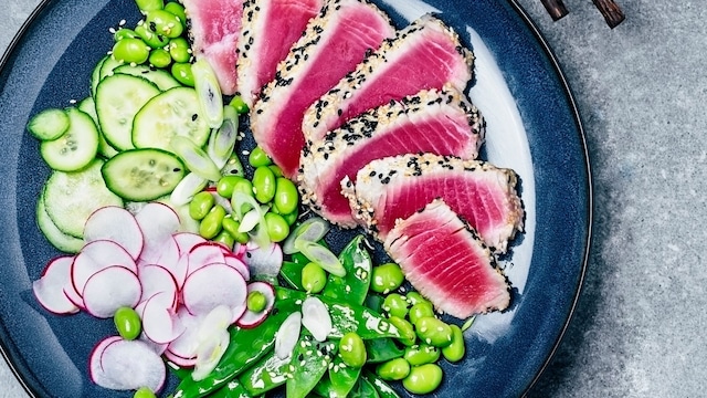 A plate of sliced tuna steak with fresh salad (radish, cucumber, edamame, and sugar snap peas) on gray background