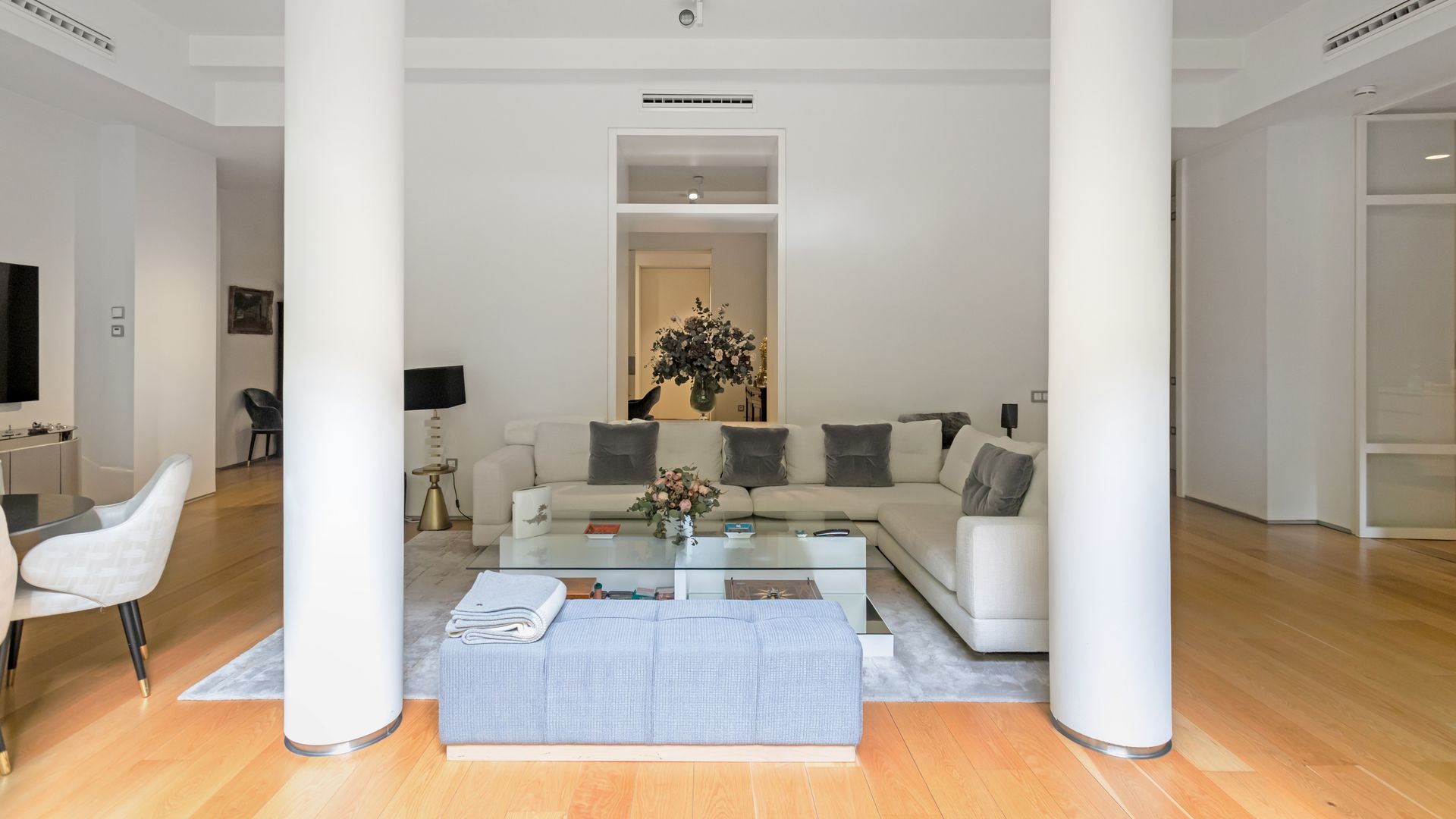 Salón con columnas, sofá blanco, cojines grises, alfombra gris, mesa de centro de cristal, puf a modo mesa 