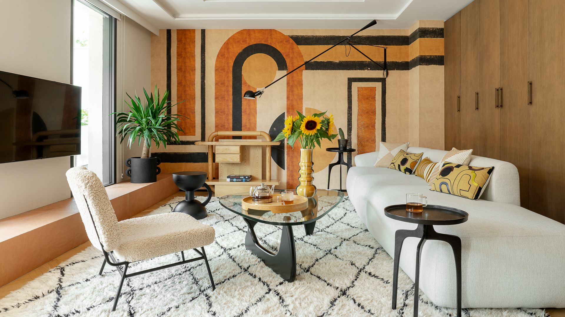 Salón con sofá blanco, pared con mural de papel pintado en colores tierra, alfombra, mesa de centro de diseño con el sobre de cristal, jarrón con girasoles, butaca, televisión colgada en la pared, techos con molduras