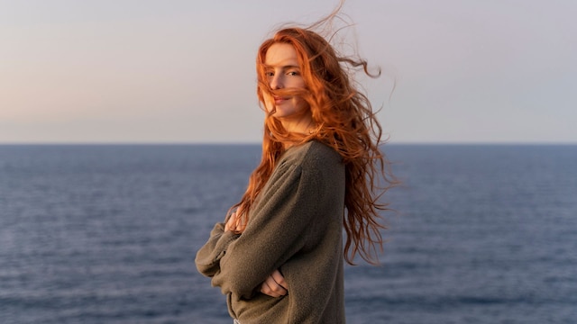 Mujer pelirroja con jersey mirando a cámara con el mar de fondo
