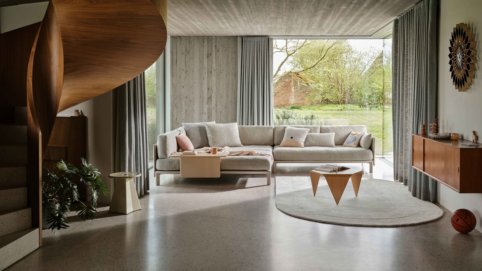 Salón moderno con techo de hormigón, gran ventanal, aparador suspendido de madera oscura, alfombra, sofá modular con mesa en un lateral