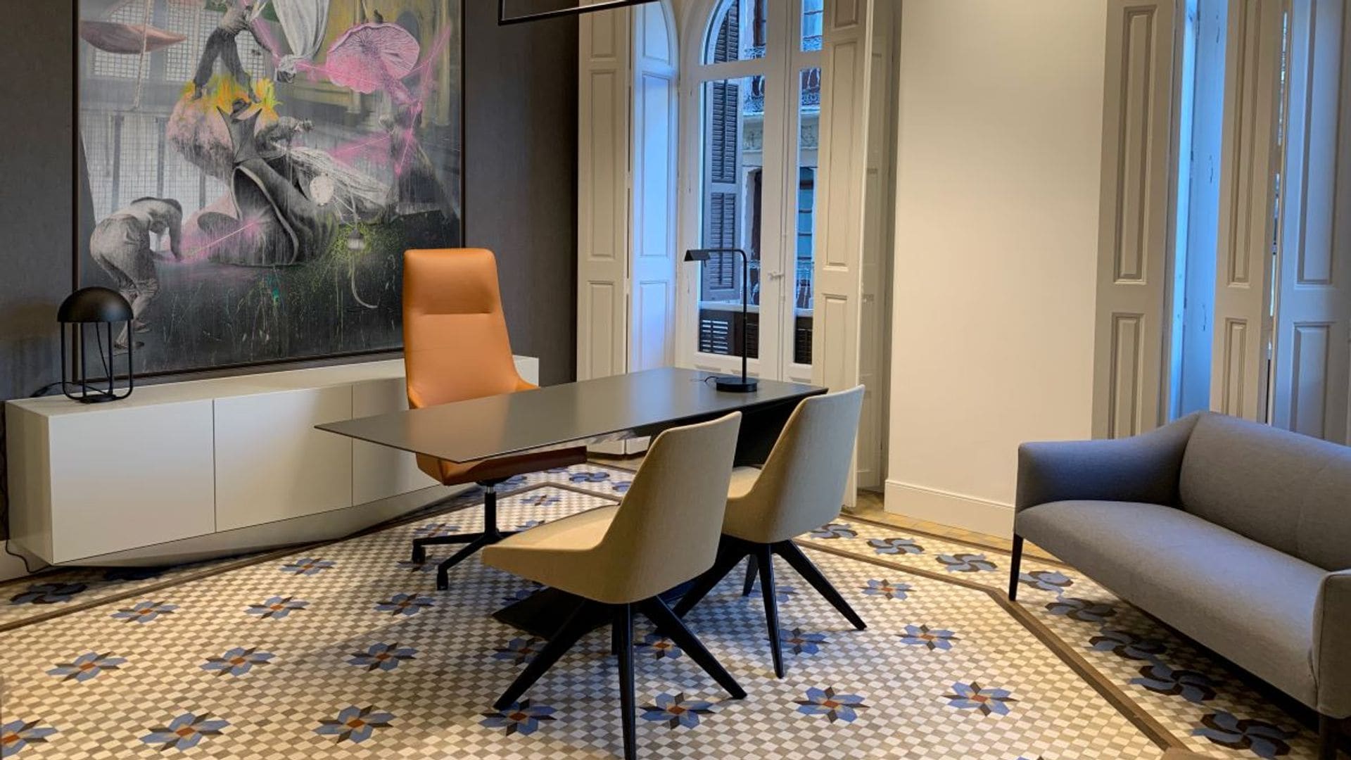 Despacho con suelo hidráulico, mesa negra, silla de oficina, sofá gris, cuadro en la pared, aparador bajo, ventanas y contraventanas de madera blanca, lámpara de techo negra, lámpara de sobremesa negra 