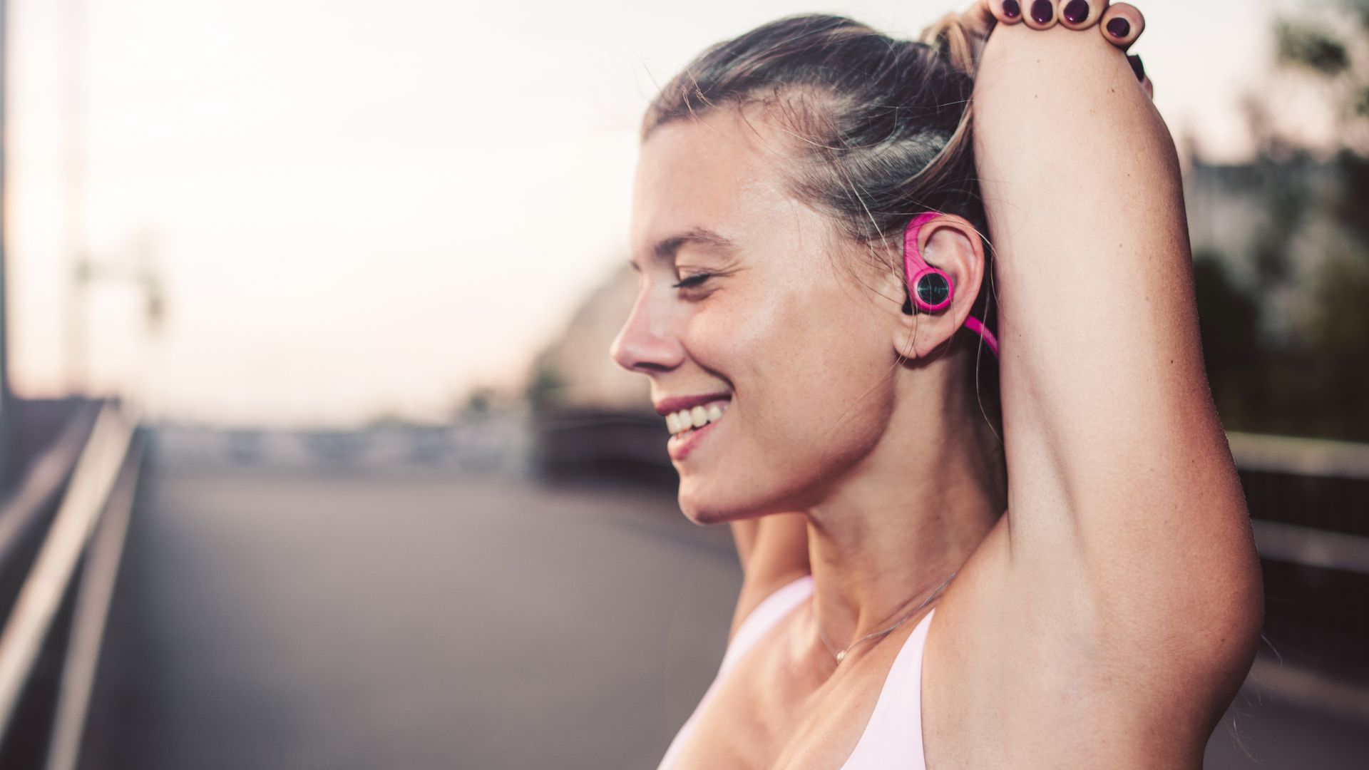 Mujer haciendo estiramientos después de salir a correr por las calles de la ciudad, con ropa de primavera-verano y auriculares inalámbricos en fucsia.