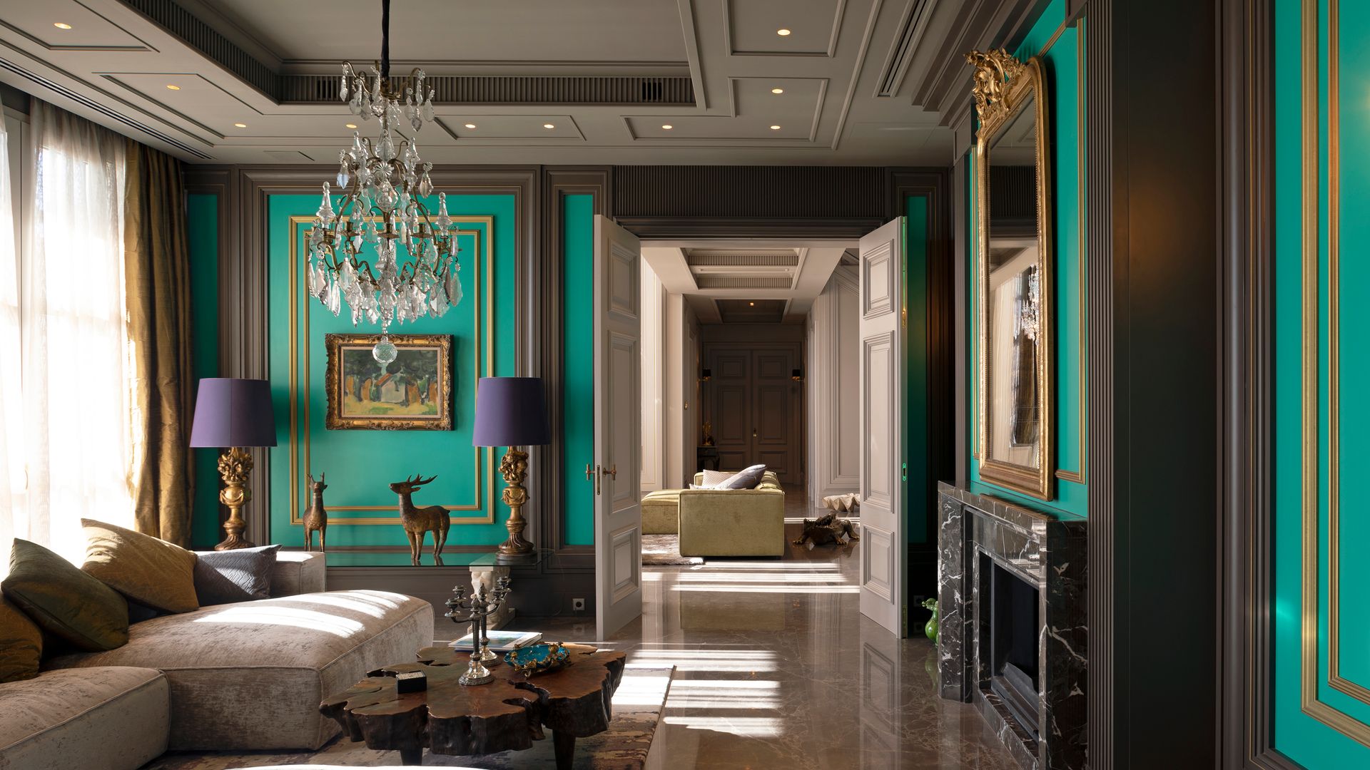 Salón con chimenea de mármol, paredes verdes con molduras, lámpara de techo de cristal, lámparas de sobremesa con pantalla morada, sofá beige, suelo de mármol