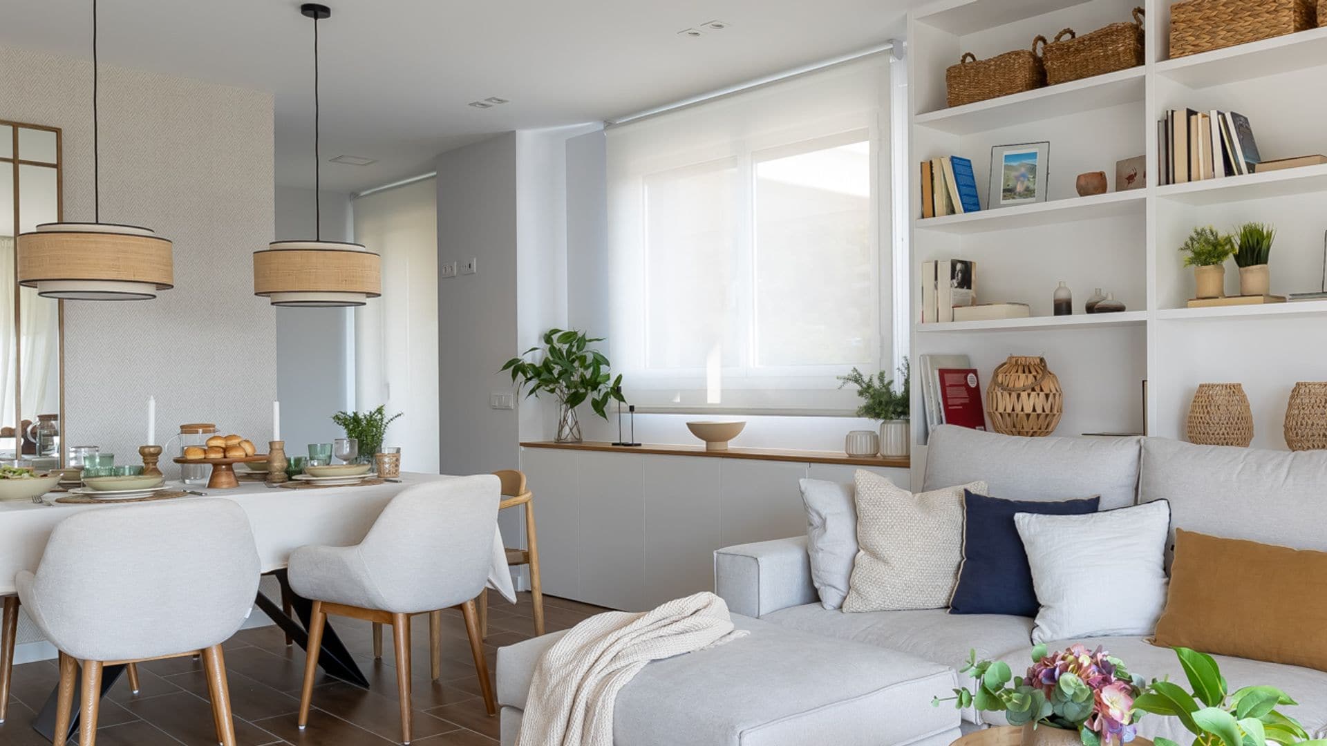 Salón comedor con estantería de obra a medida blanca, sofá blanco, mesa de centro de madera con el pie metálico negro, espejo, mesa de comedor con mantel blanco, lámparas de techo de fibras vegetales, aparador bajo la ventana, estor