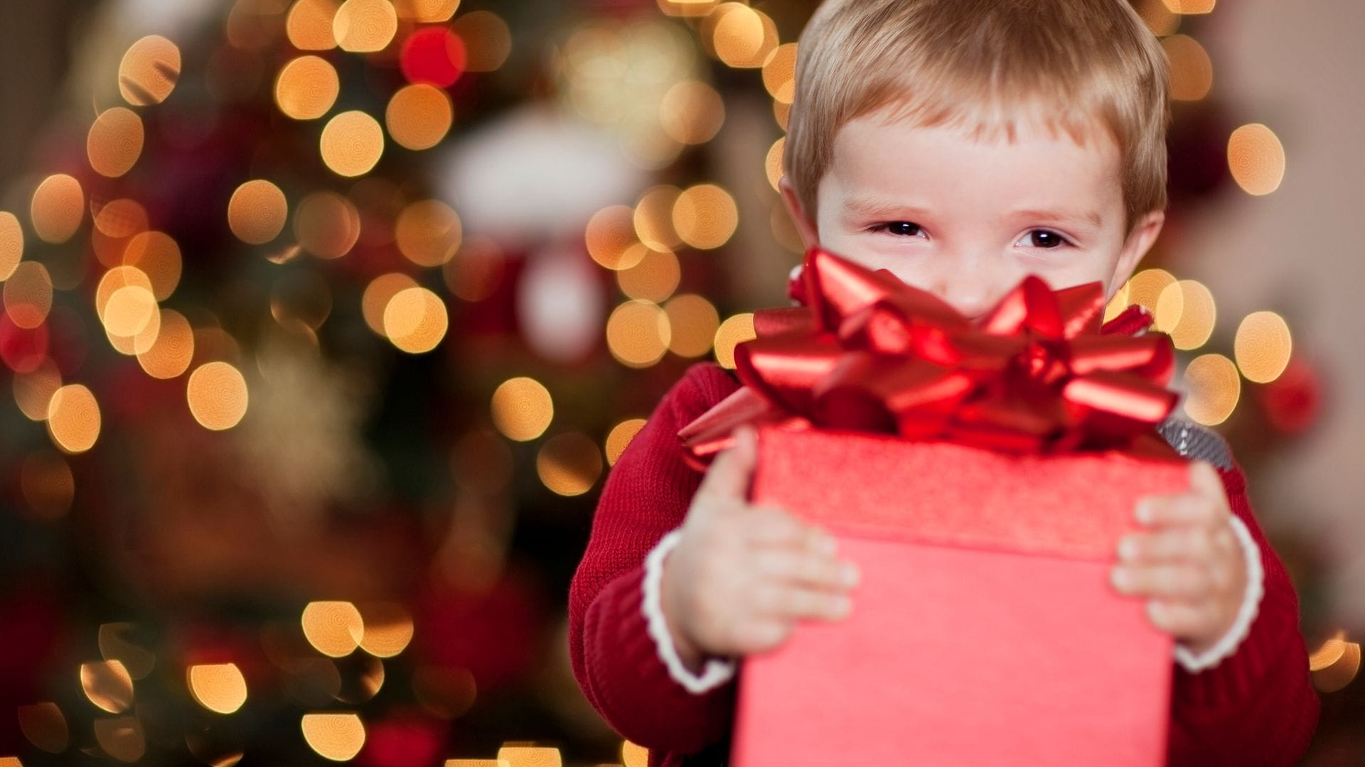 Niño sonriendo detrás de una caja de regalo roja, que tiene con las manos. El fondo está iluminado por Navidad.