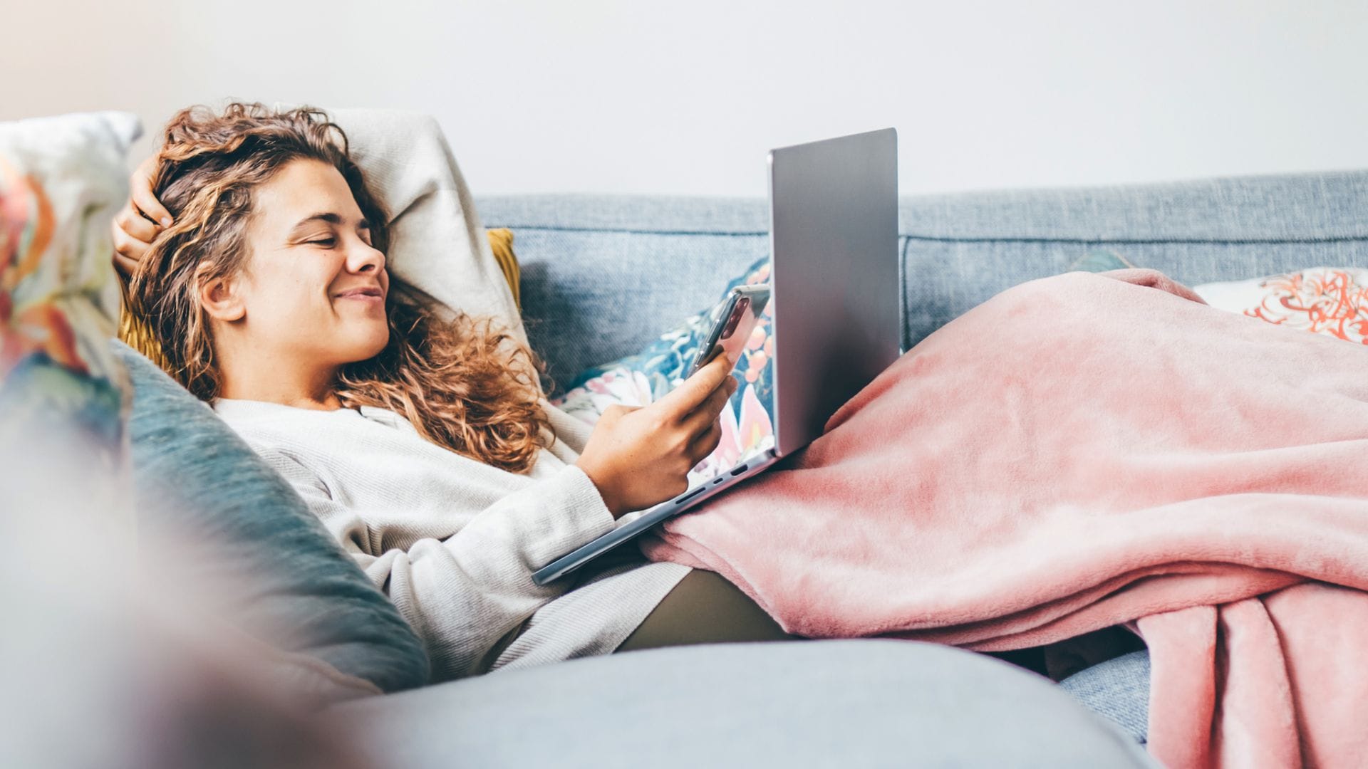 Chica joven tumbada en el sofá del salón con una manta rosa encima, muy abrigada y sonriente, con un ordenador portátil y un teléfono móvil en las manos.