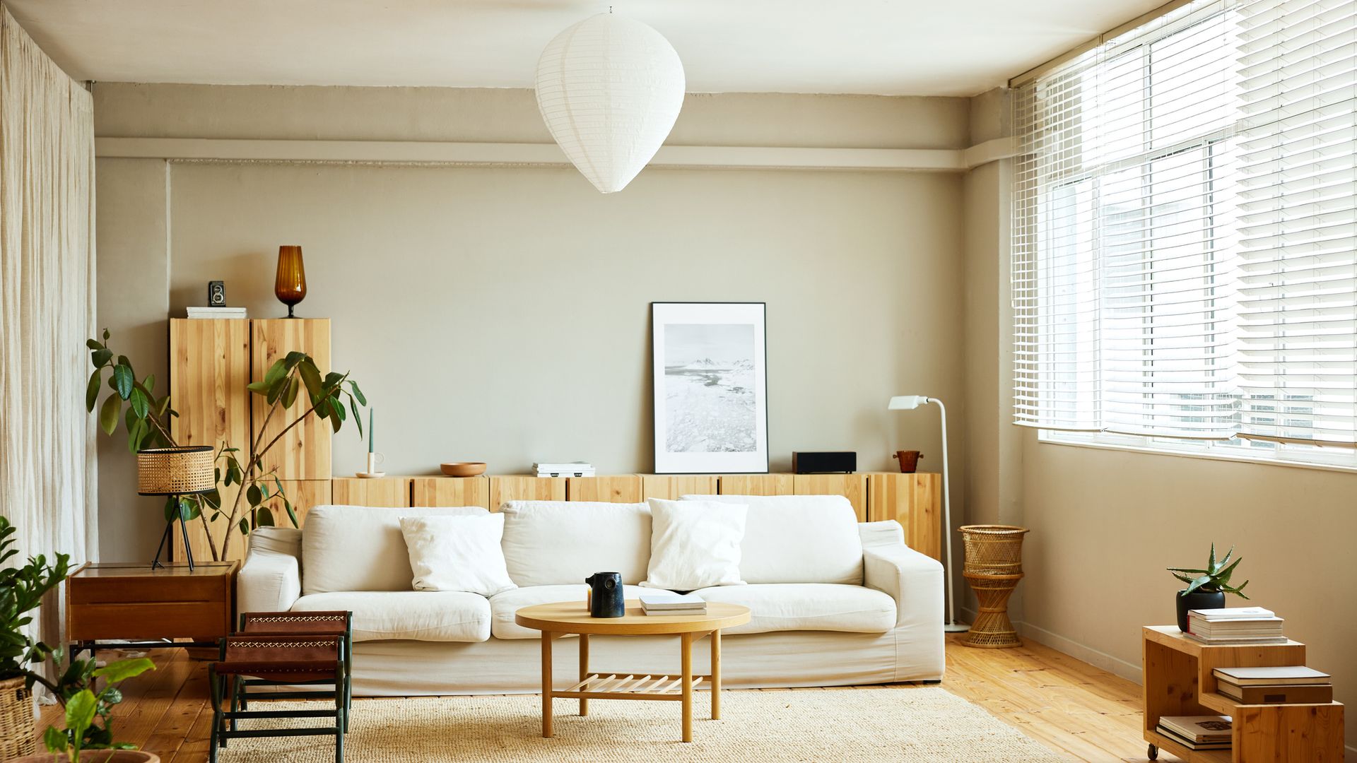 Salón decorado en blanco, 'beige' y madera con plantas. Iluminado, con un sofá y módulos de almacenaje al fondo.