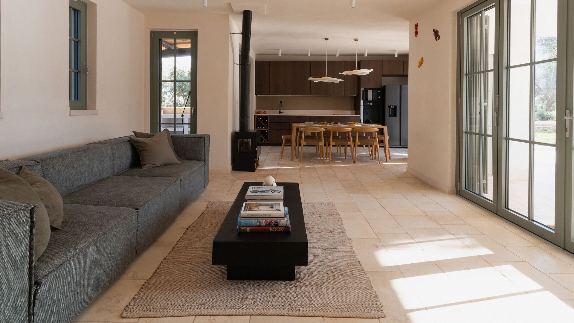Cocina abierta al salón con chimenea, sofá gris y mesa de centro negra, ventanas verdes con salida al jardín 