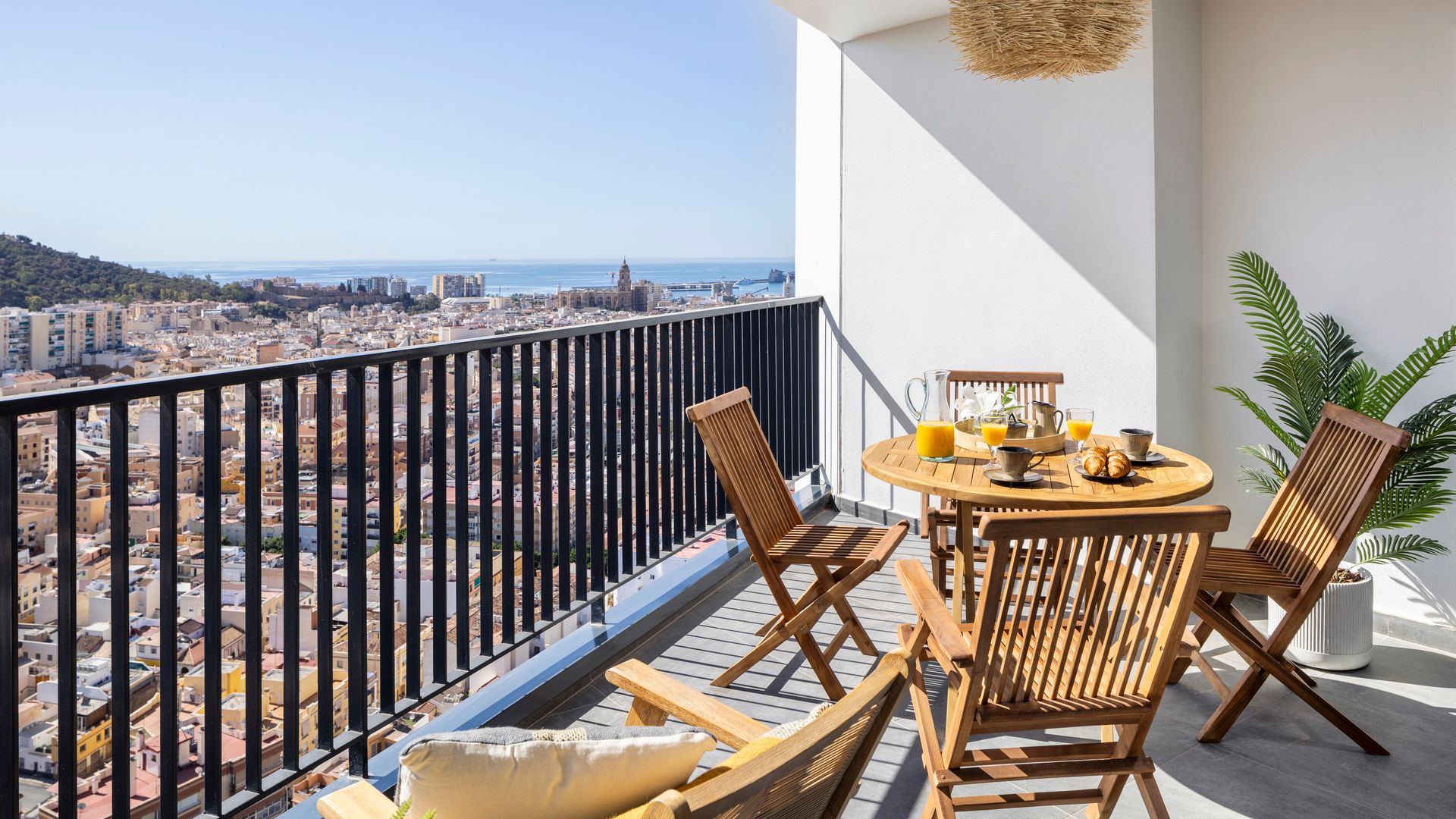 Terraza con barandilla negra, mesa de comedor redonda, sillas de madera plegables, lámpara de techo de fibras vegetales 