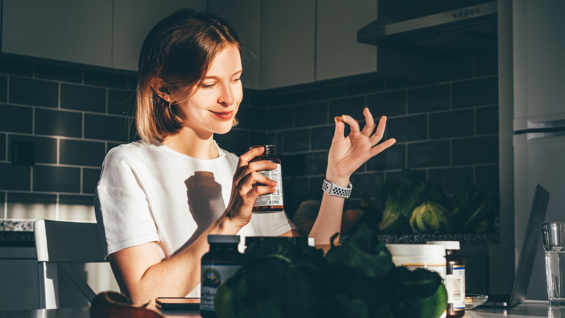 Mujer joven mirando una pastilla de suplemento alimenticio en la cocina.