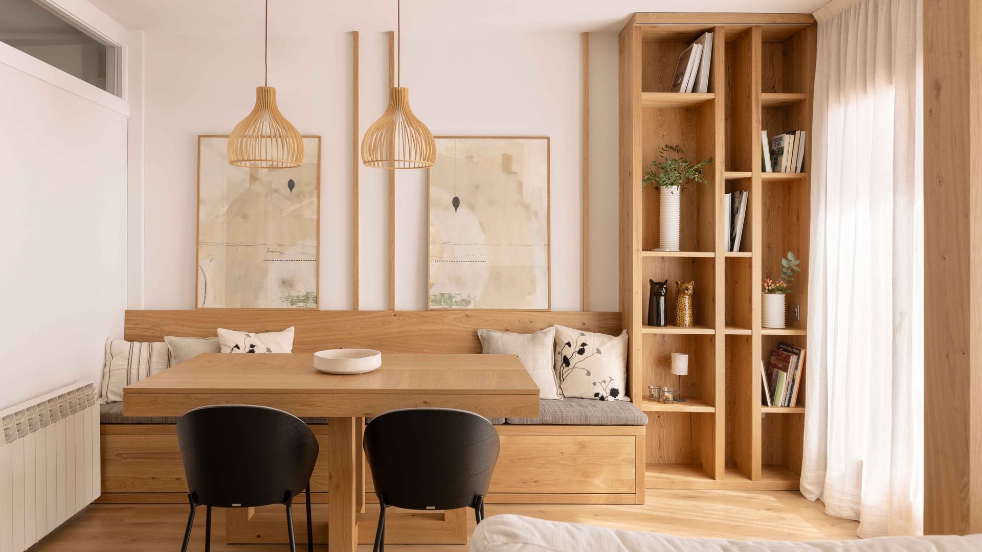 Salón comedor de estilo japandi, con mesa y banco de madera, librería de madera, lámpara de fibras vegetales, sillas negras, cuadros en la pared, cortinas blancas