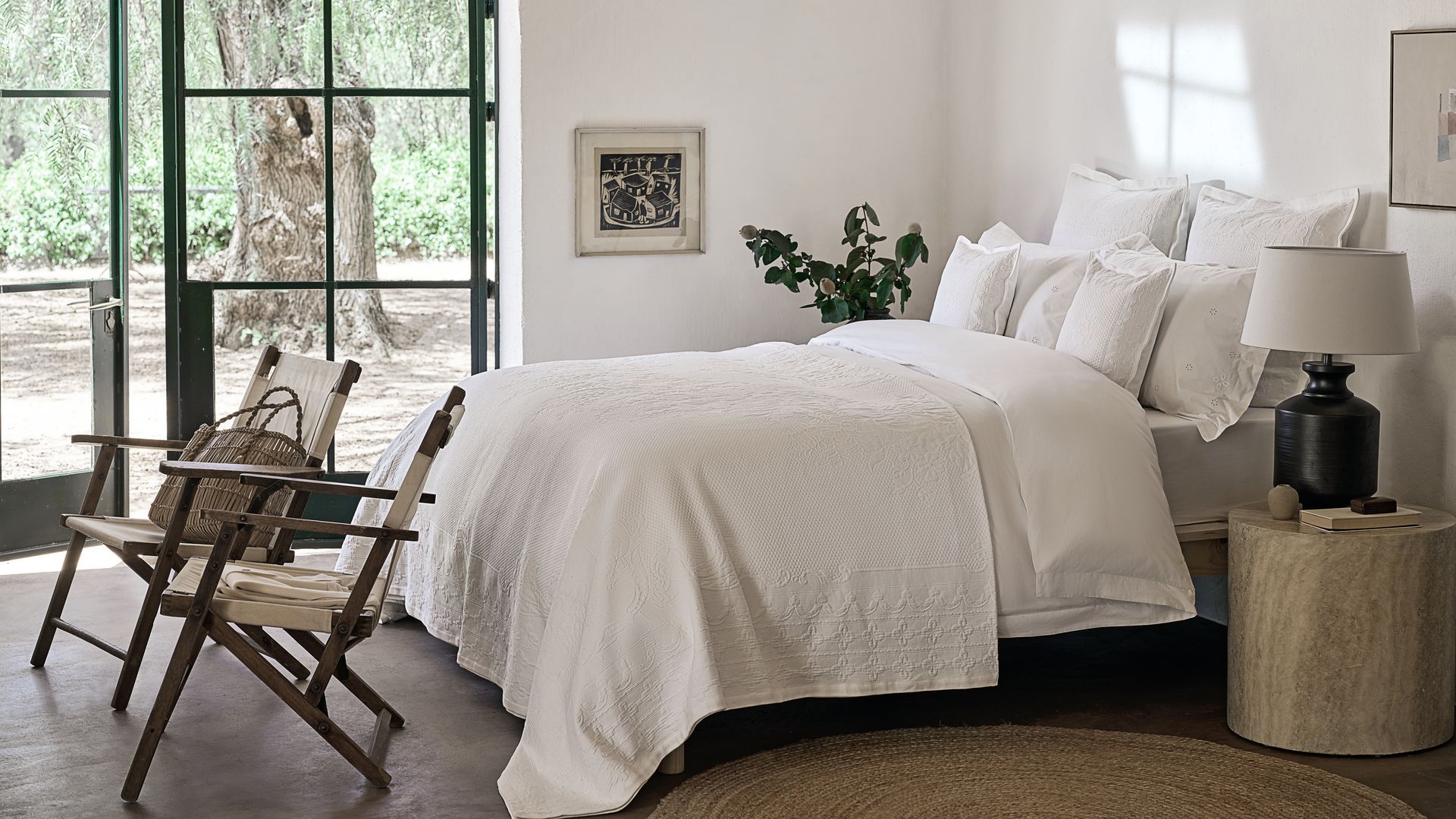 Dormitorio blanco, con salida al jardín, sillas a los pies de la cama, sábanas blancas, mesita de noche, almohadas, lámpara de sobremesa negra