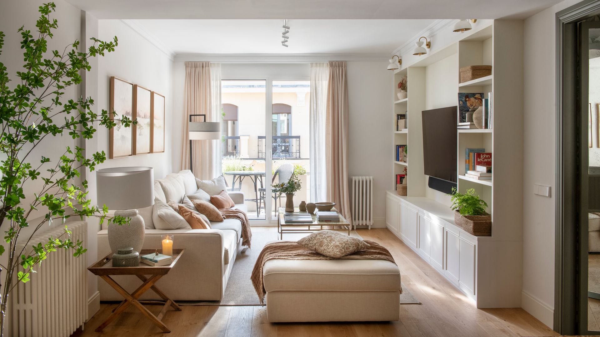 Salón con alfombra, cortinas en las ventanas, sofá beiges, suelo de madera, librería a medida blanca, mesa auxiliar de madera tipo tijera