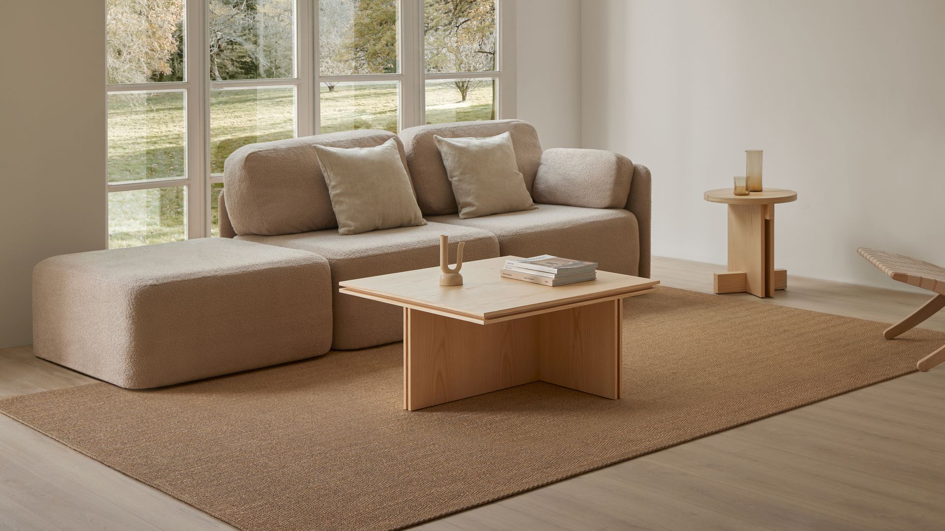 Salón minimalista con sofá modular beige, mesa de centro de madera, ventana con cuarterones, mesa auxiliar de madera clara y alfombra de fibras vegetales