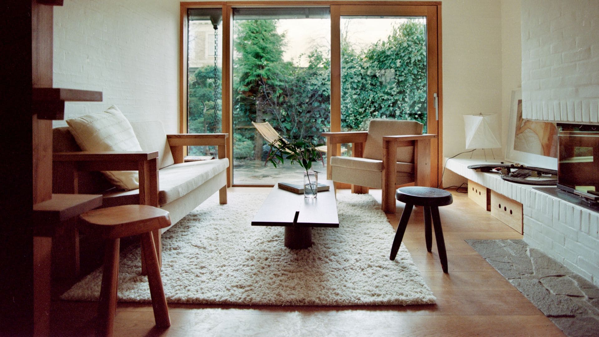 Salón con chimenea, alfombra de lana de pelo largo, sofá con estructura de madera y asiento blanco, salida al jardín con ventanas de madera, suelo de barro