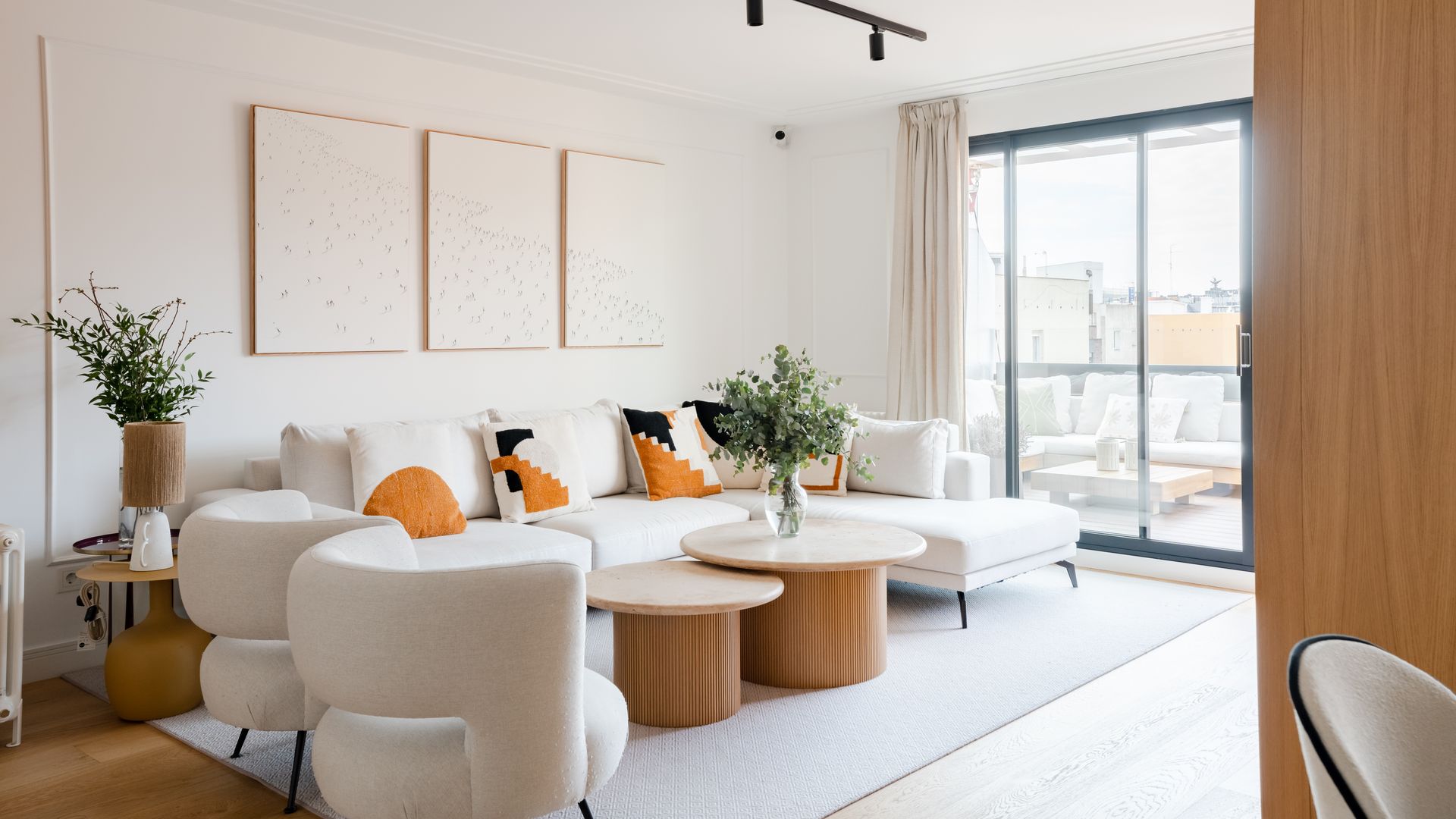 Salón con sofá blanco con chaise longue, butacas blancas, alfombra blanca, ventanal con salida terraza, jarrones con ramas verdes, cuadros en la pared del sofá, mesas de centro de madera 