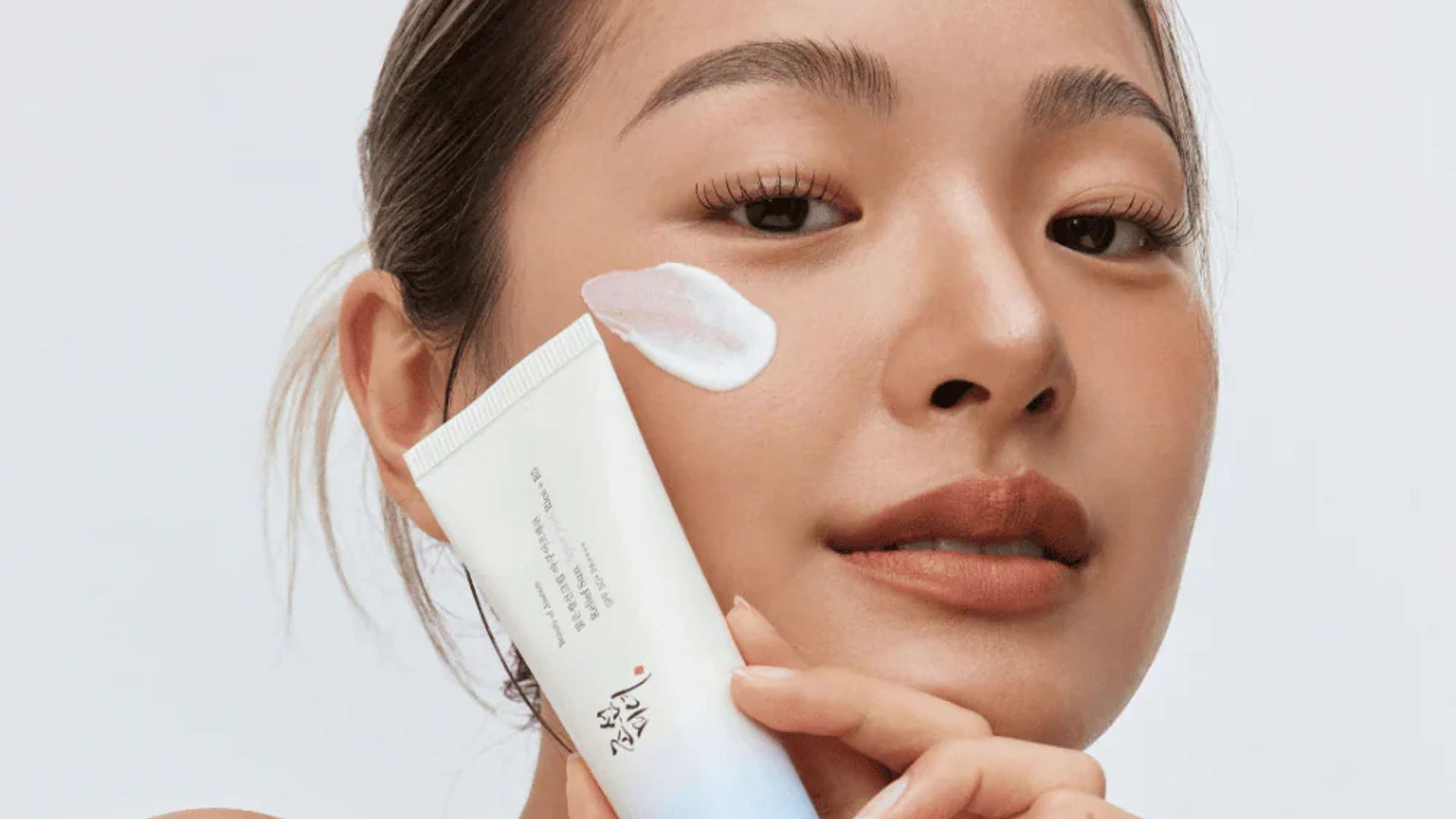 Chica joven asiática con una crema solar coreana entre las manos, mirando hacia la cámara en primer plano.