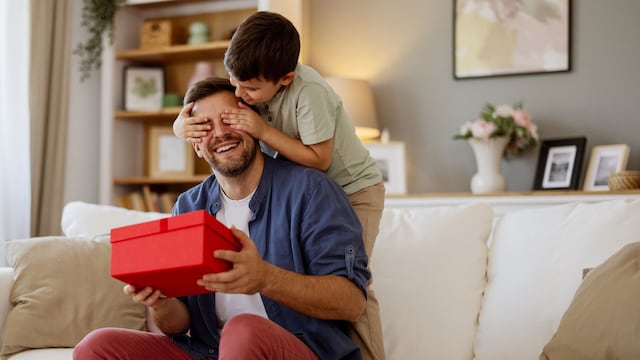 Día del Padre. Niño dándole un regalo a su padre en el sofá de su casa mientras le tapa los ojos para que no vea cuál es la sorpresa