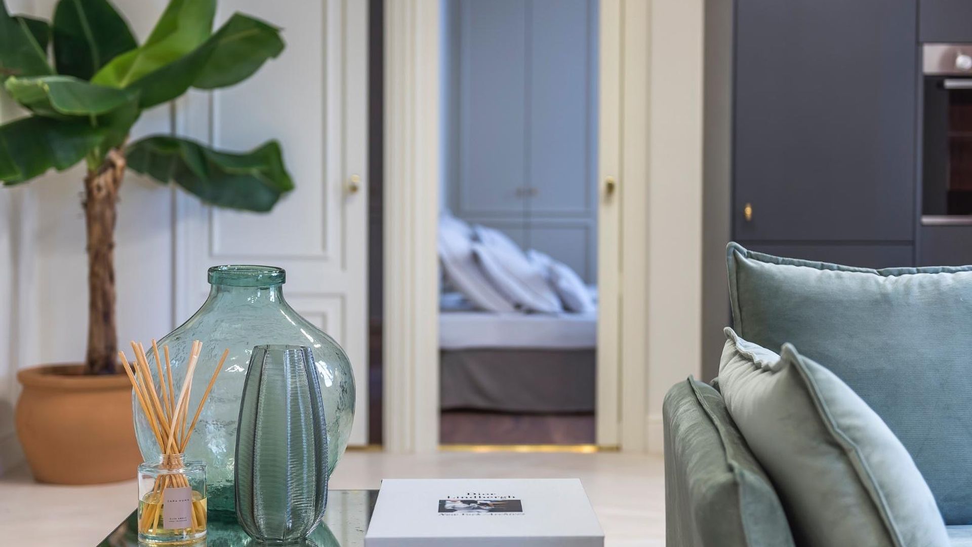 Cocina abierta al salón, con vistas del dormitorio, con muebles de cocina azules oscuros, sofás en gris azulado, jarrones de cristal verde, ficus XL en el suelo con maceta de barro, ambientador mikado