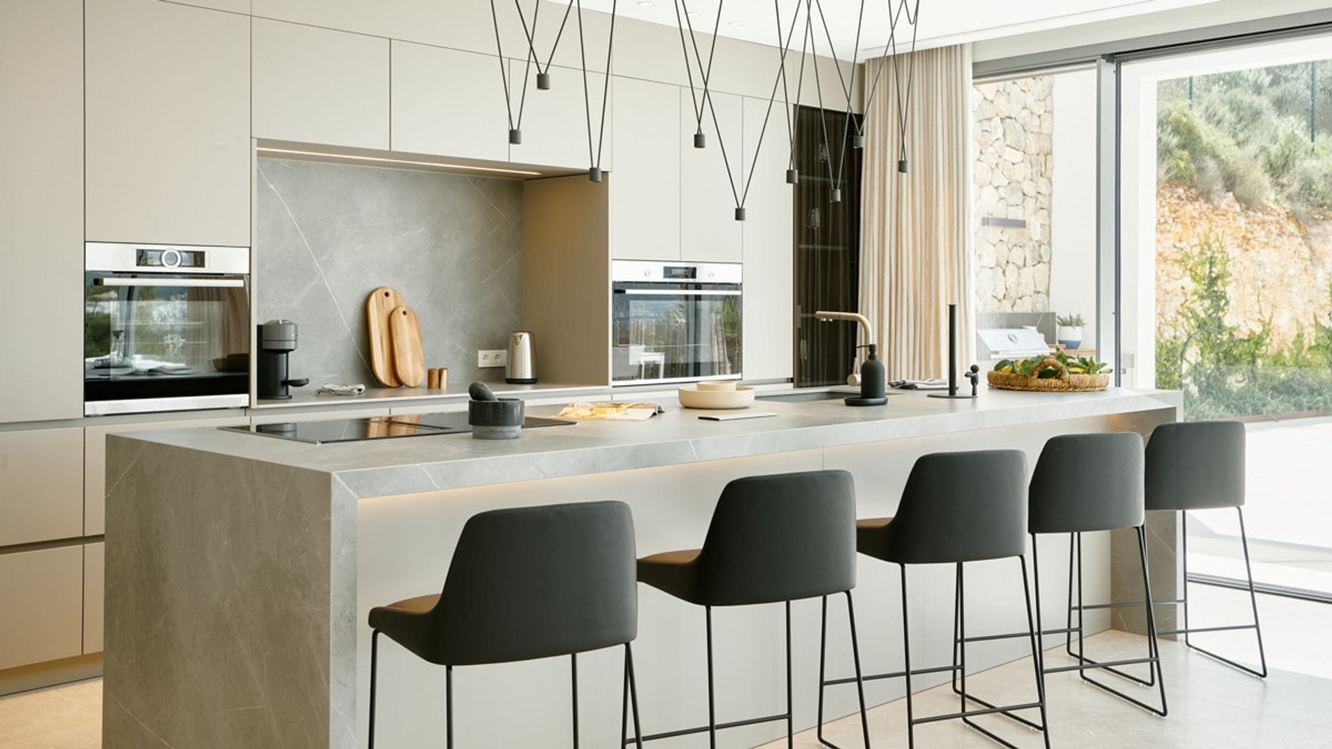 Cocina abierta con isla con salida al jardín, muebles de color crema, lámpara de techo, taburetes negros