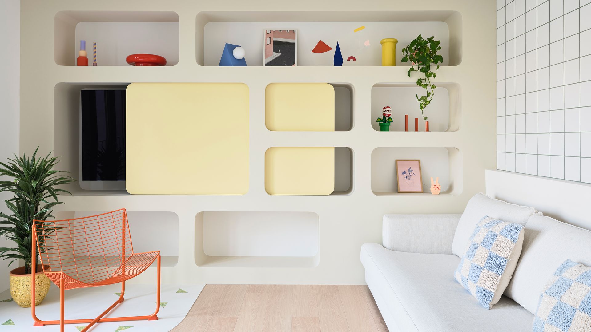 Salón con estantería a medida, televisión, sofá blanco, cojines azules y blancos, adornos colores, silla metálica naranja, plantas