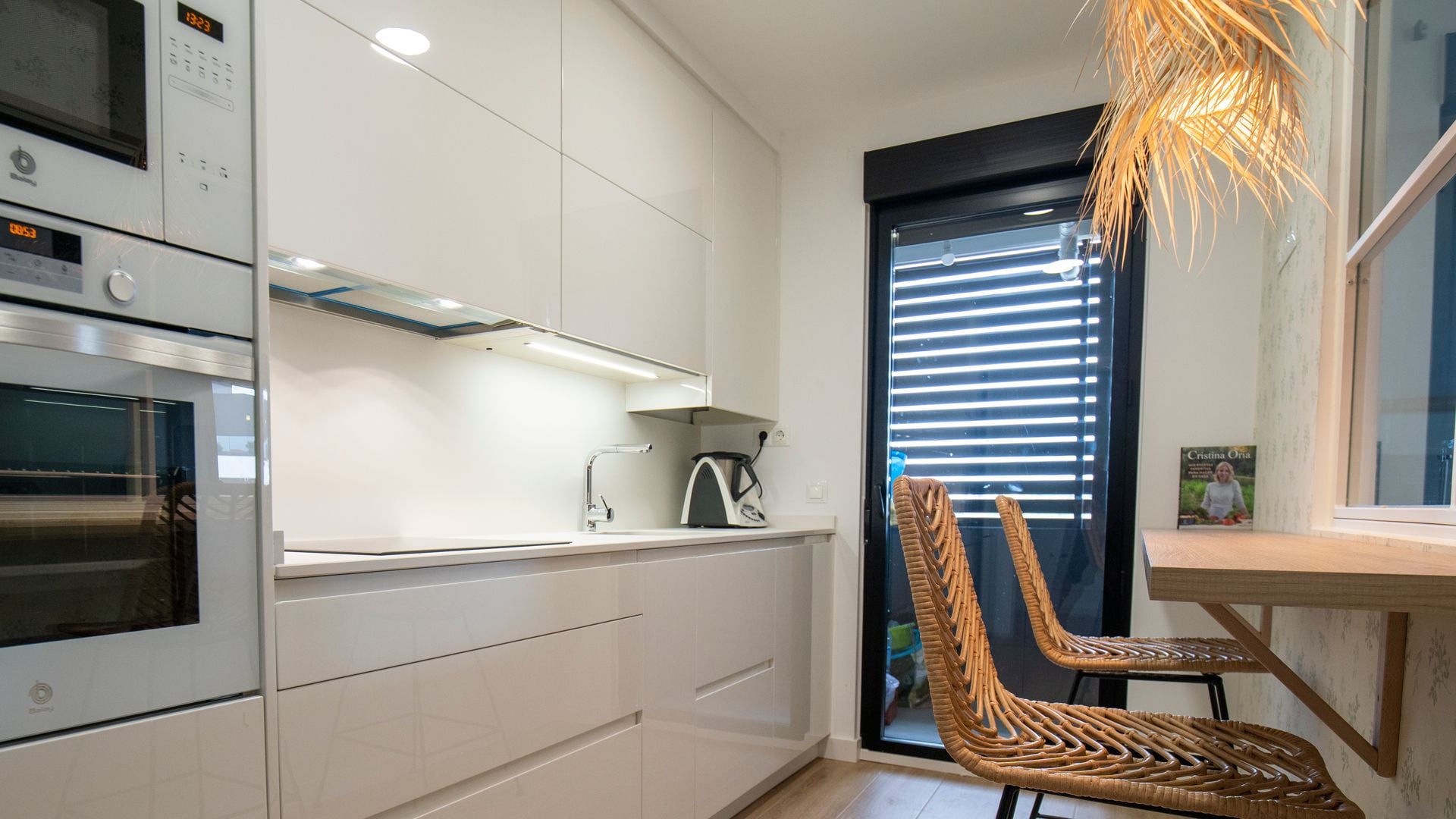 Cocina blanca con muebles hasta el techo, horno y microondas de acero en columna, barra de madera, taburetes y lámpara de techo de fibras naturales