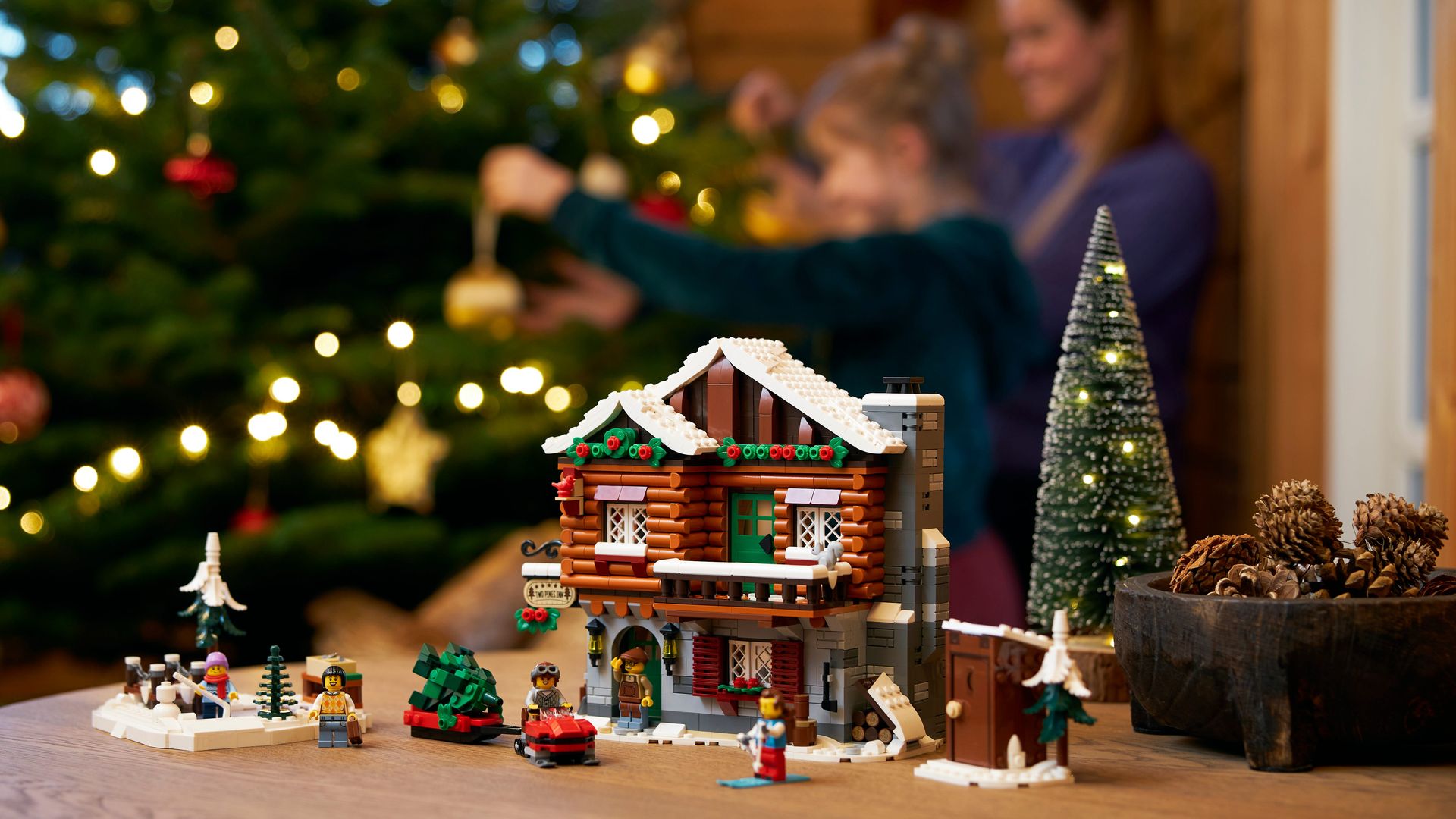 LEGO Casa Alpina de Navidad sobre una mesa mientras decoran por detrás un árbol dos niñas sonrientes.