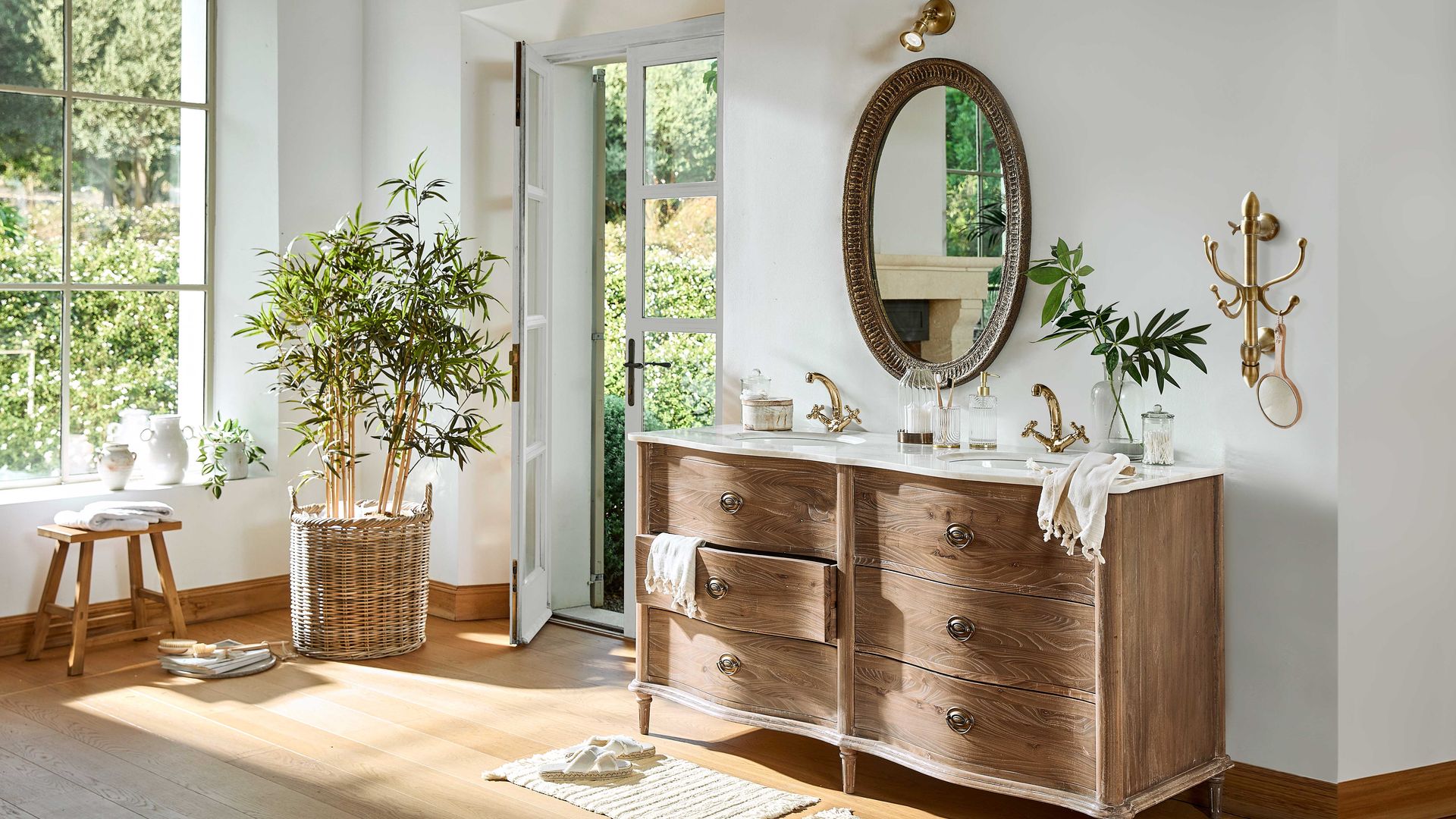Baño XL con gran ventanal, suelo de madera, mueble de madera, espejo, planta en cesto, elementos dorados