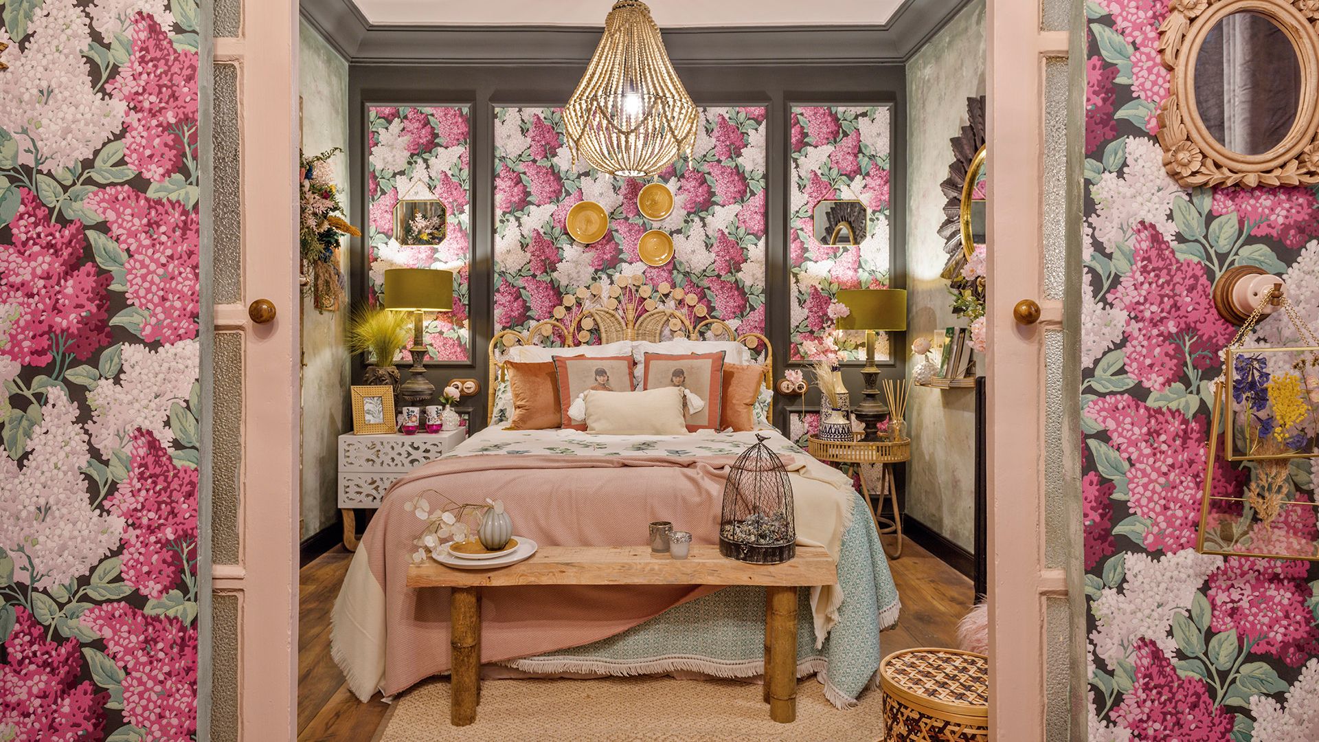 Dormitorio con papel pintado de flores rosas y blancas, molduras verdes, banco de madera, cojines en la cama, lámpara de techo de bambú