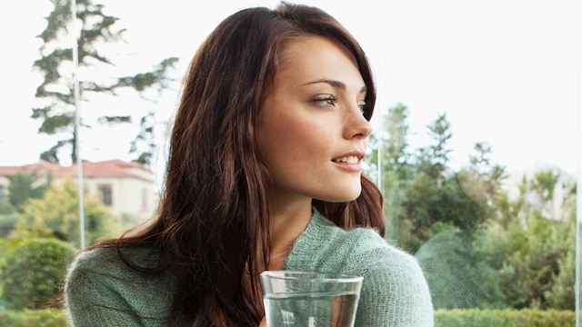 Mujer morena con jersey verde sentada en una ventana con un vaso de agua