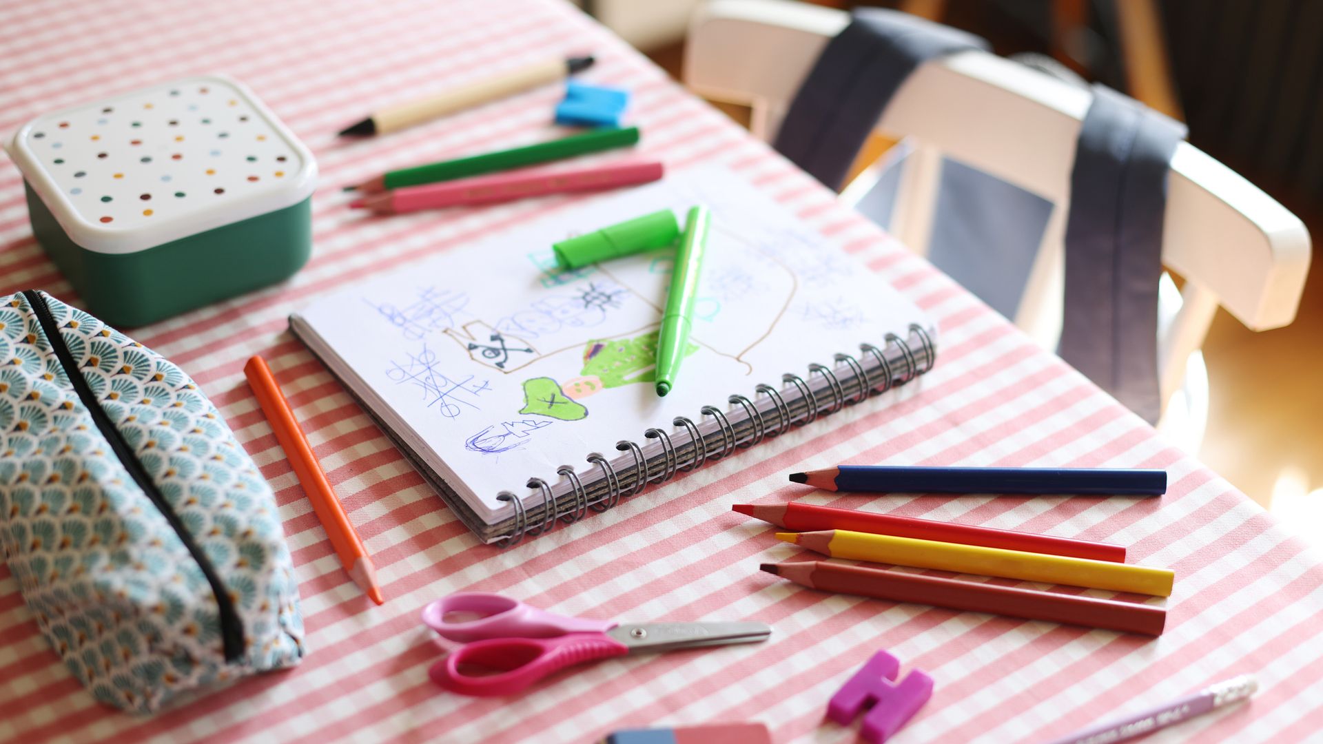 Material escolar repartido en una mesa, con un estuche cerrado, una mochila colgando de la silla y un táper.