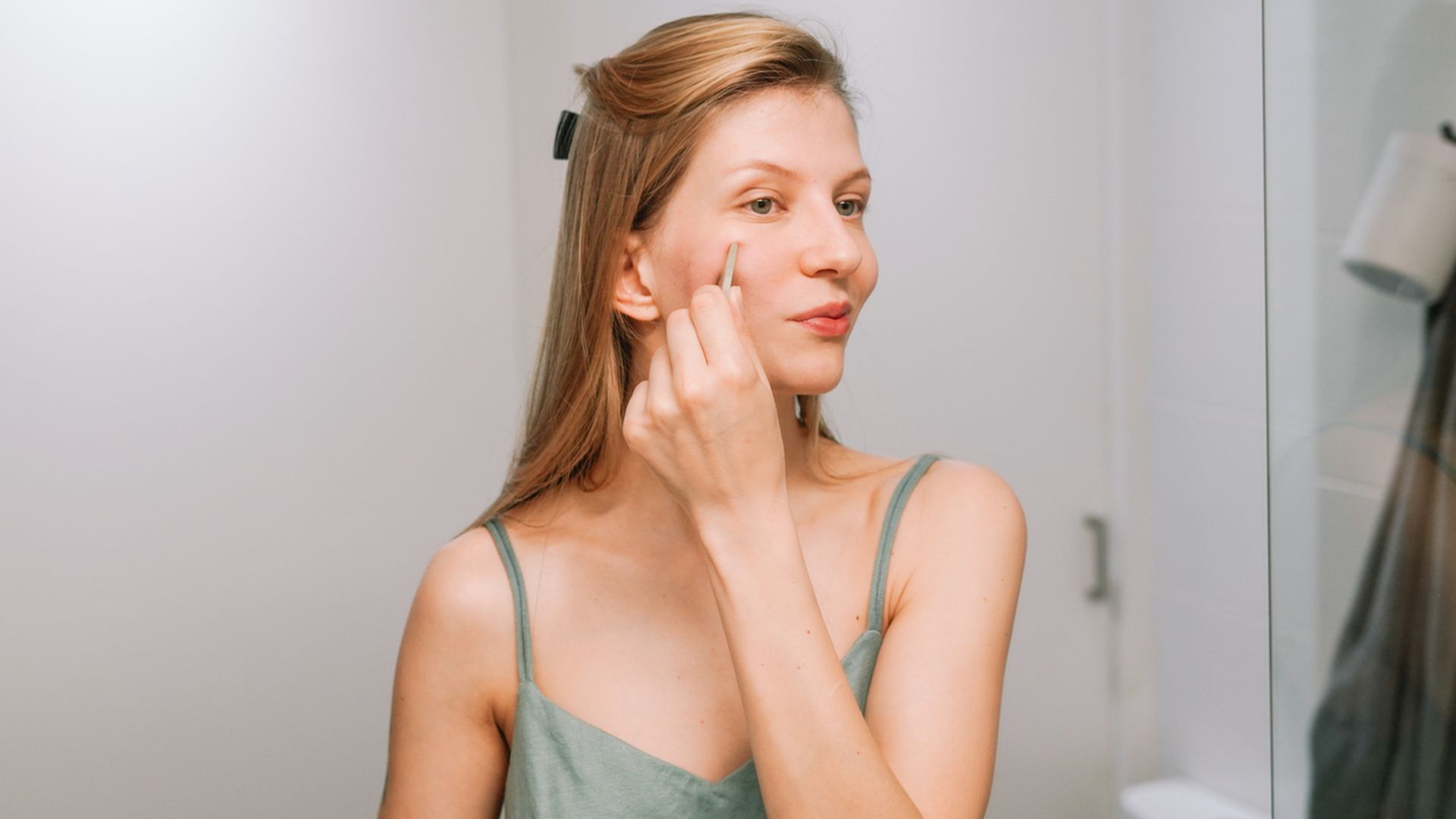 Mujer joven rubia con piel lisa y clara aplicándose productos de belleza en la mejilla en su cuarto de baño